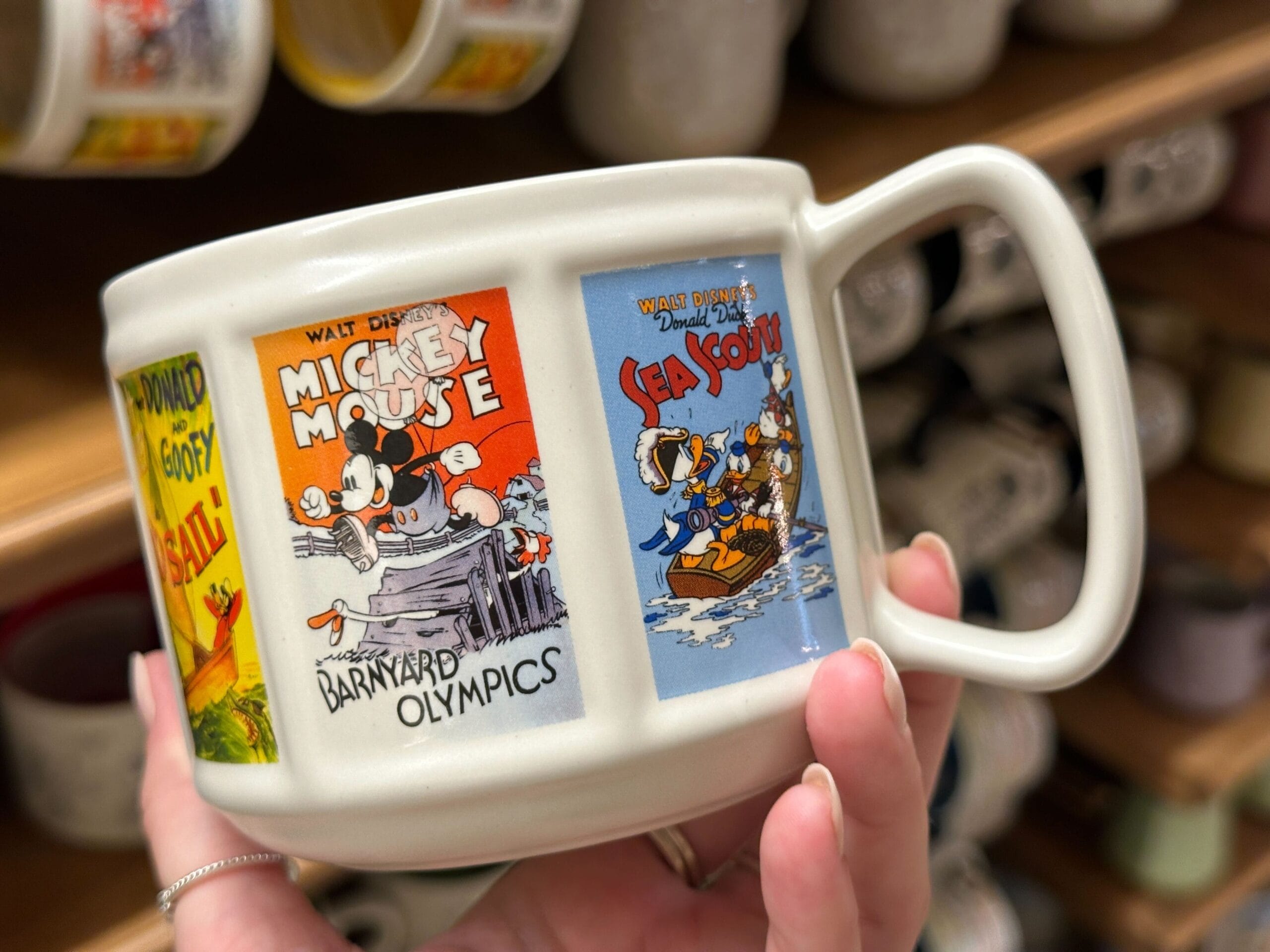 A hand holds a mug featuring vintage Mickey Mouse, Minnie, and Donald Duck comic designs.