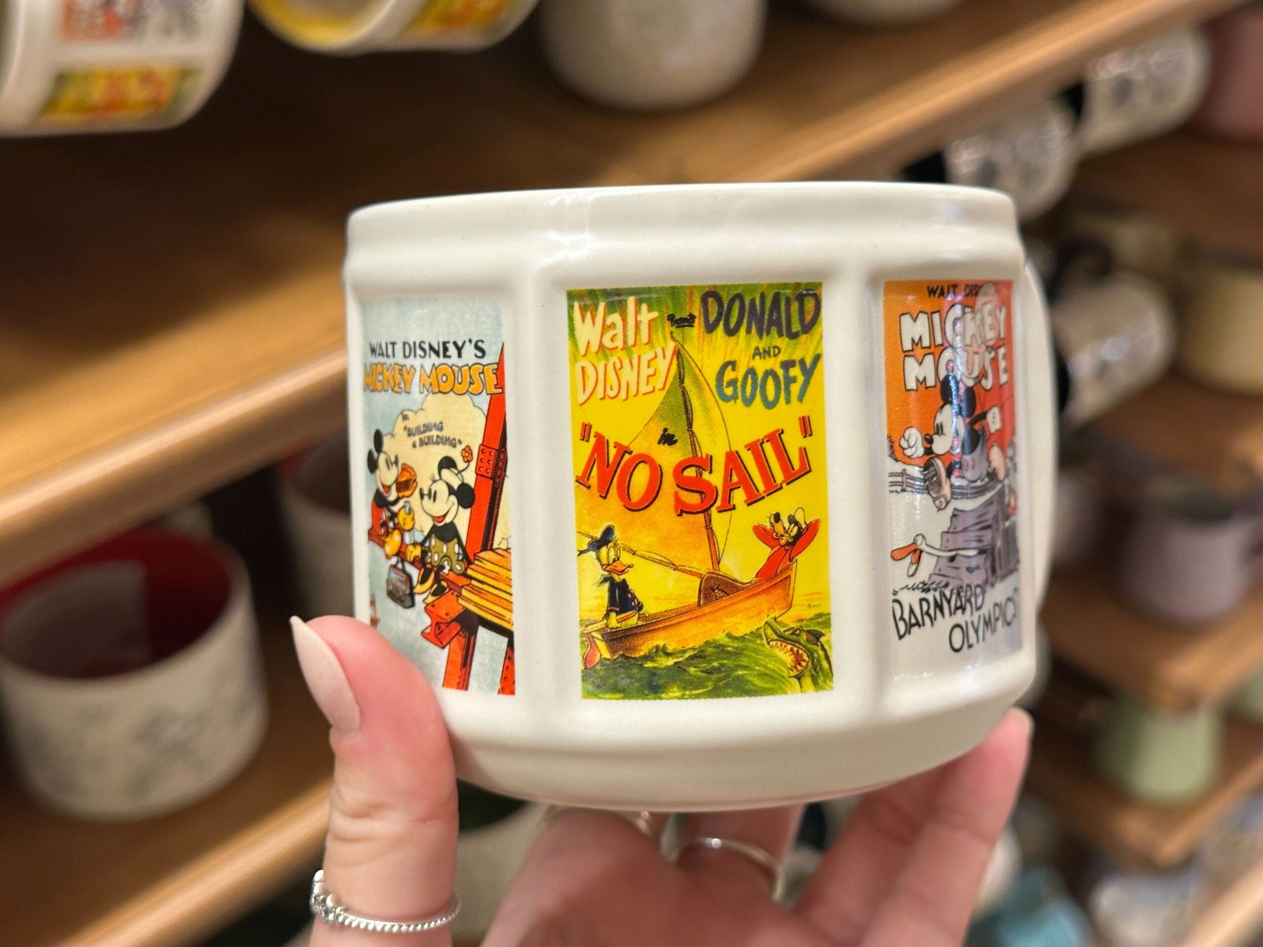 A hand holds a ceramic cup decorated with vintage Disney posters featuring Mickey, Minnie, Donald, and Goofy.