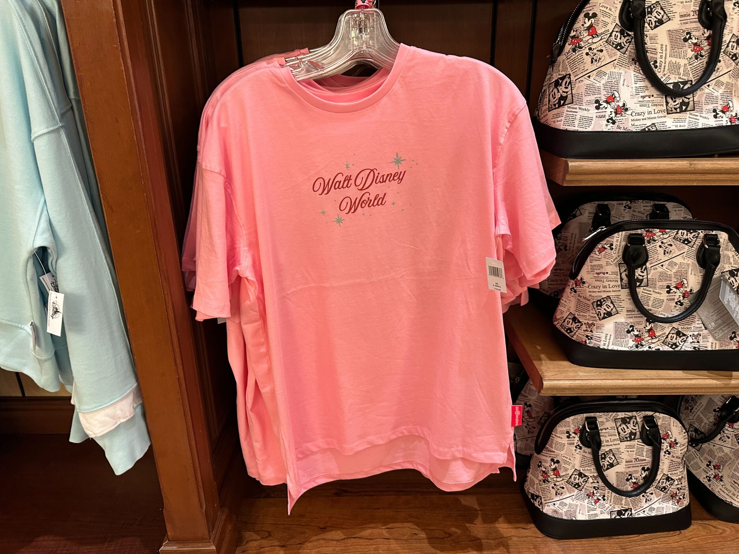 A pink t-shirt with "Walt Disney World" text hangs on a rack next to Disney-themed bags.