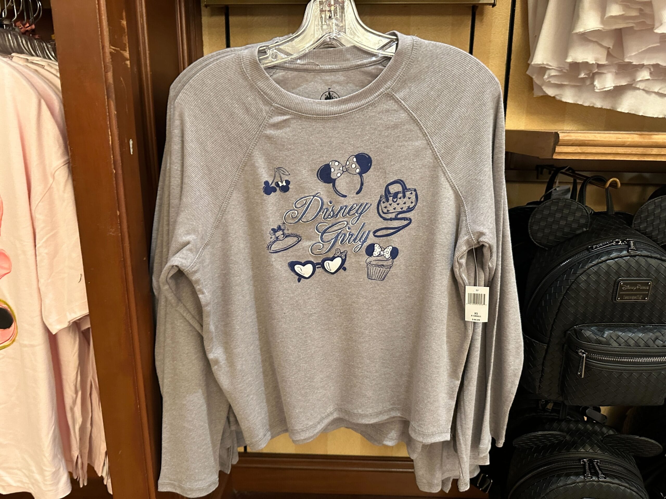 Gray long-sleeve shirt with "Disney Girly" and blue Disney-themed icons, hanging on a rack in a store.