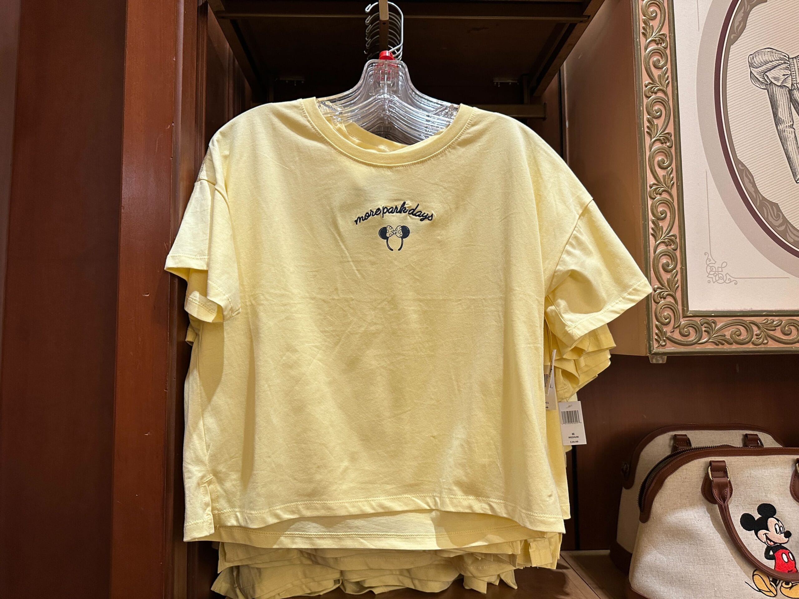 Yellow T-shirt with a small Mickey Mouse head and "mouse park days" text displayed on a hanger in a store.