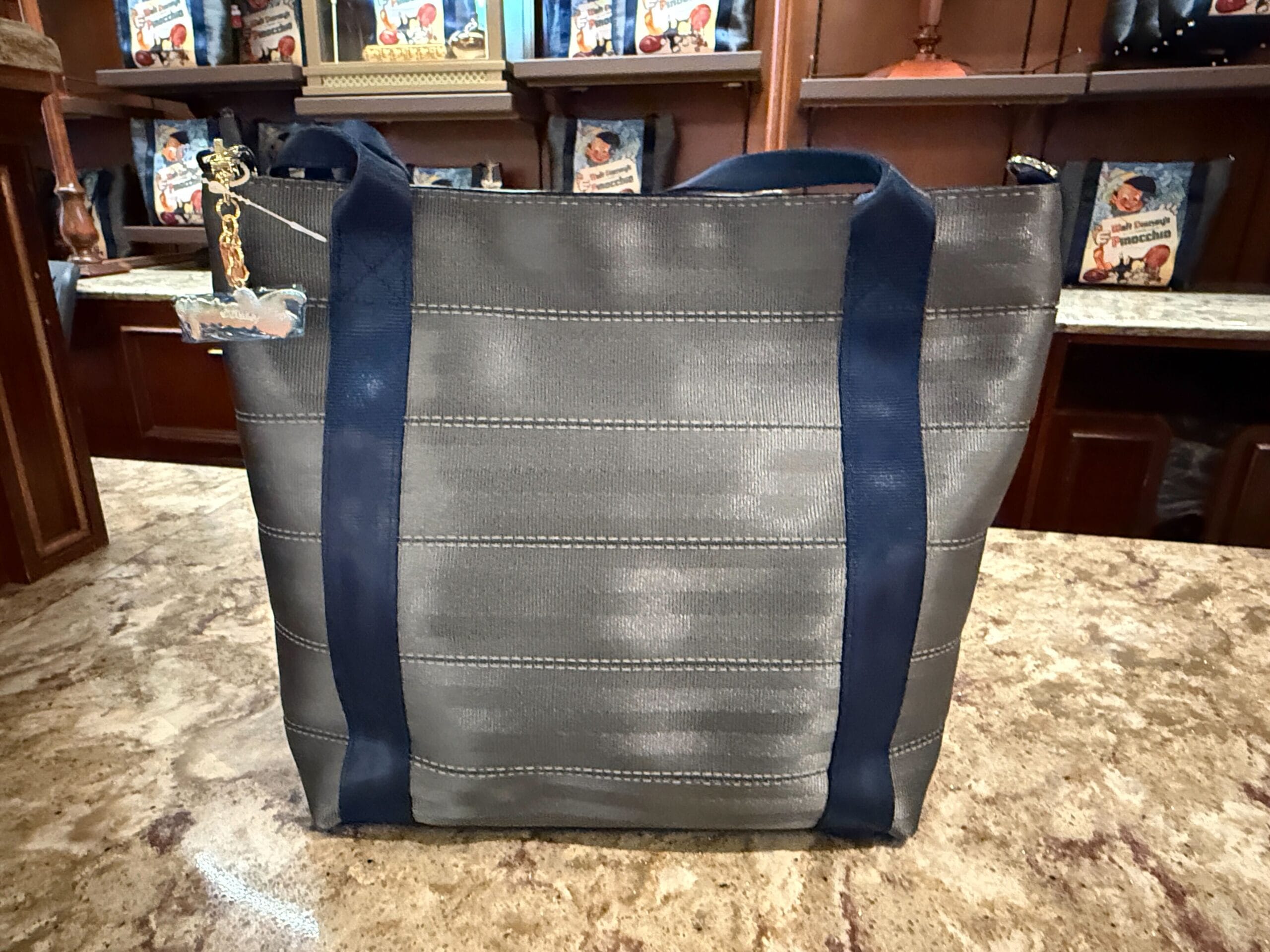 A gray tote bag with dark blue handles sits on a marble countertop in front of wooden shelves with vintage decor.