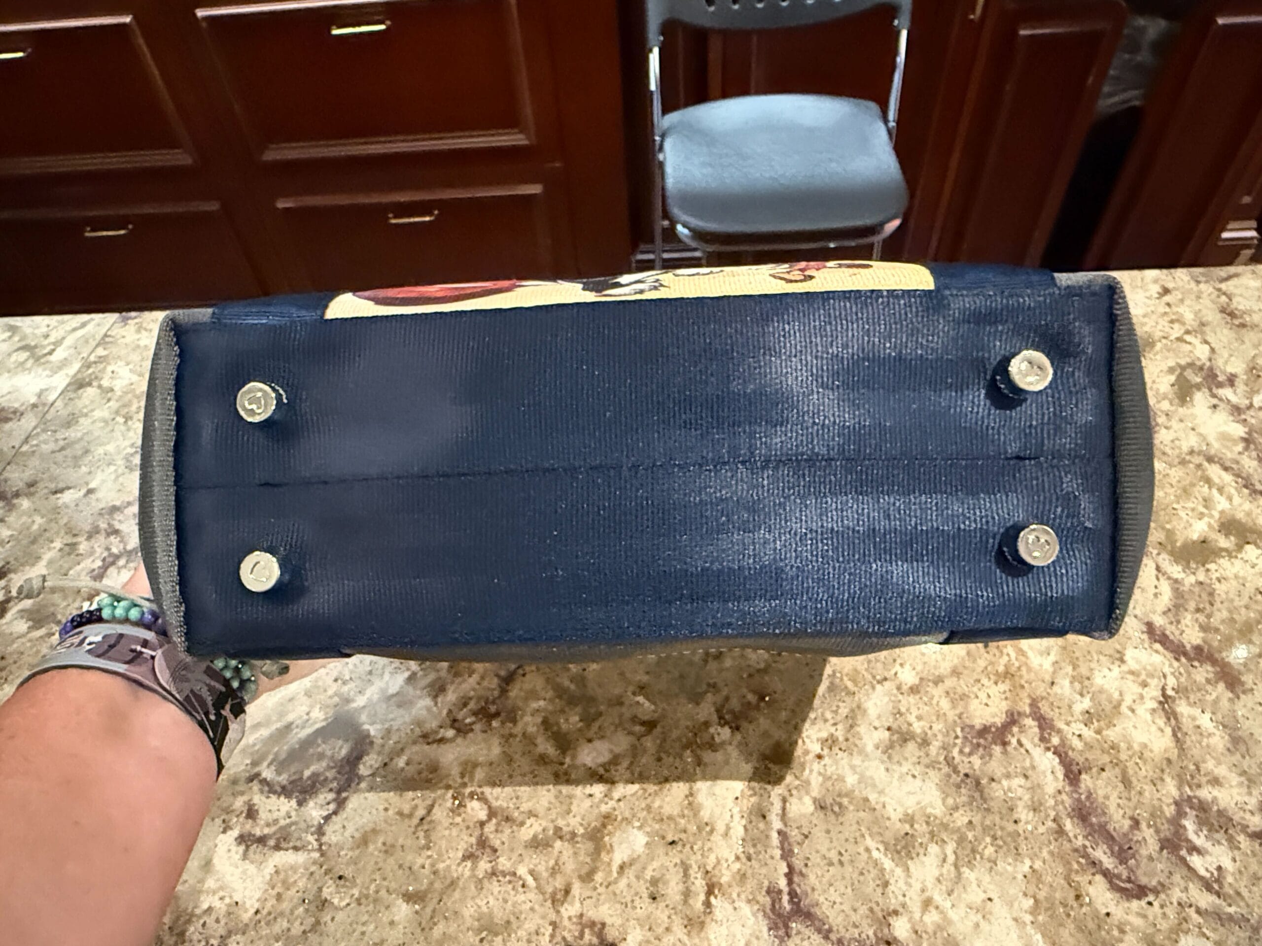 The bottom of a navy blue handbag with four metal feet, held over a marble countertop.