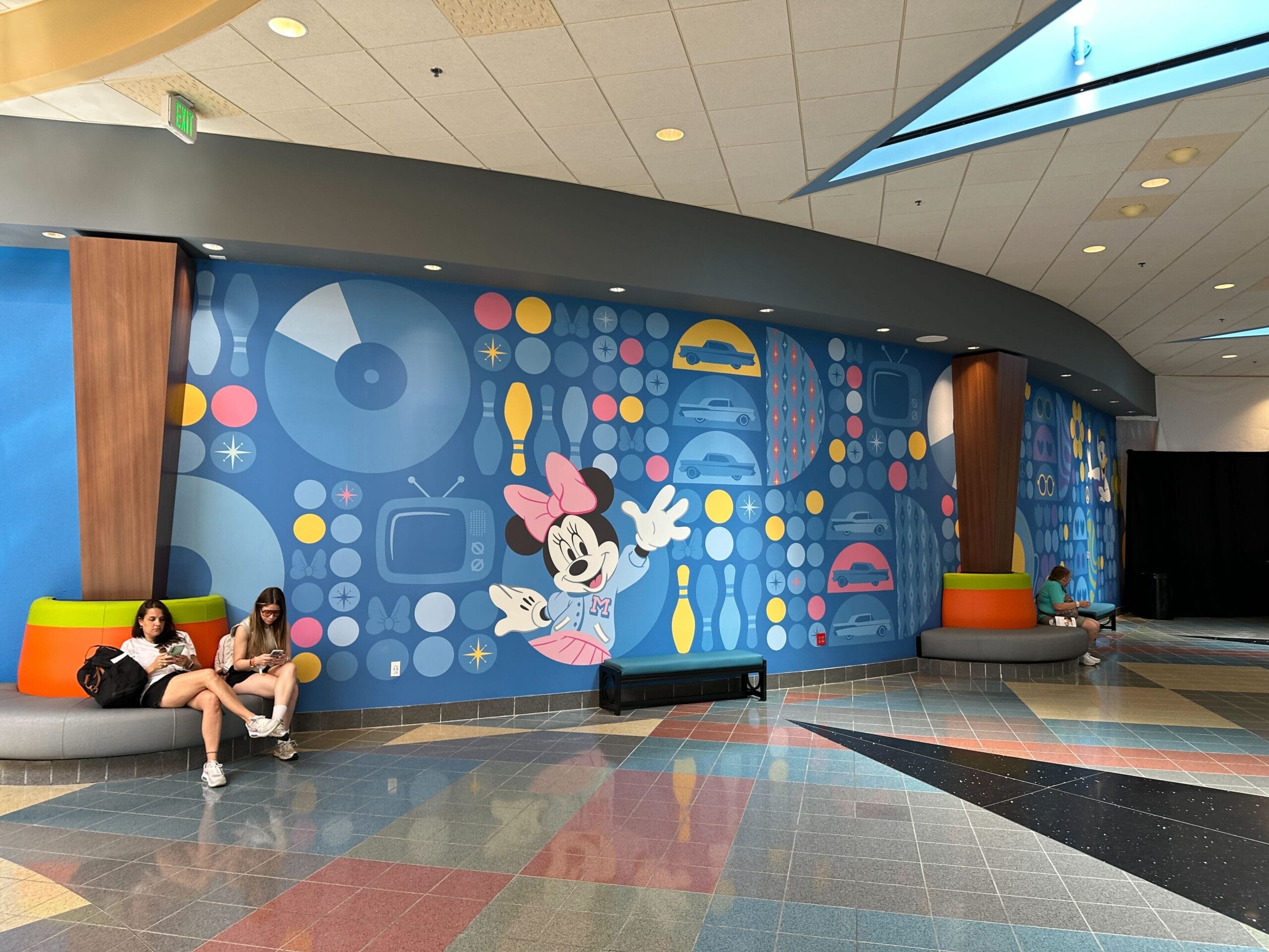 A colorful mural with Minnie Mouse, retro icons, and people sitting on modern benches in a bright indoor space.