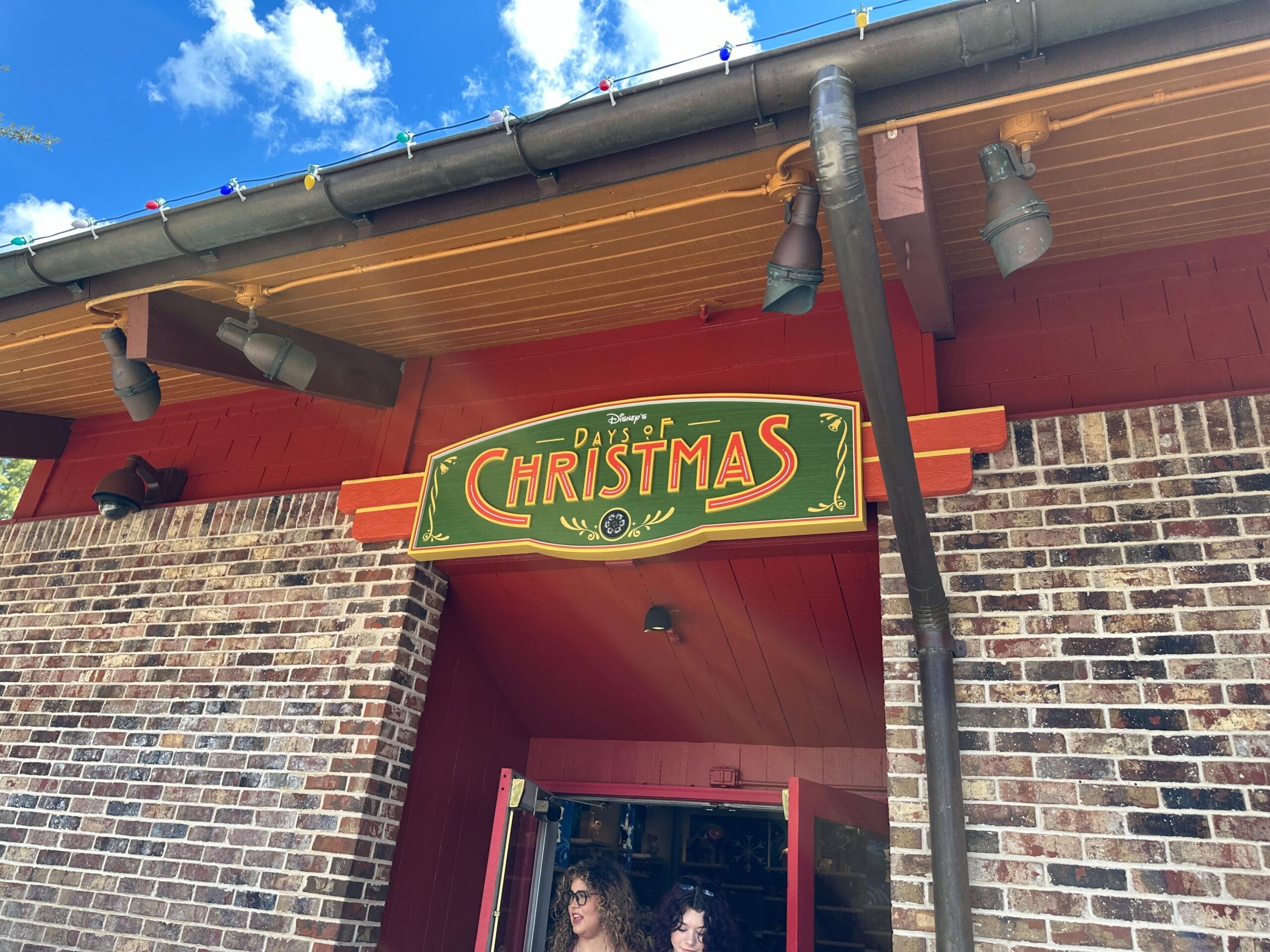 A sign reading "Days of Christmas" hangs above a red and brick building entrance under a blue sky with clouds.