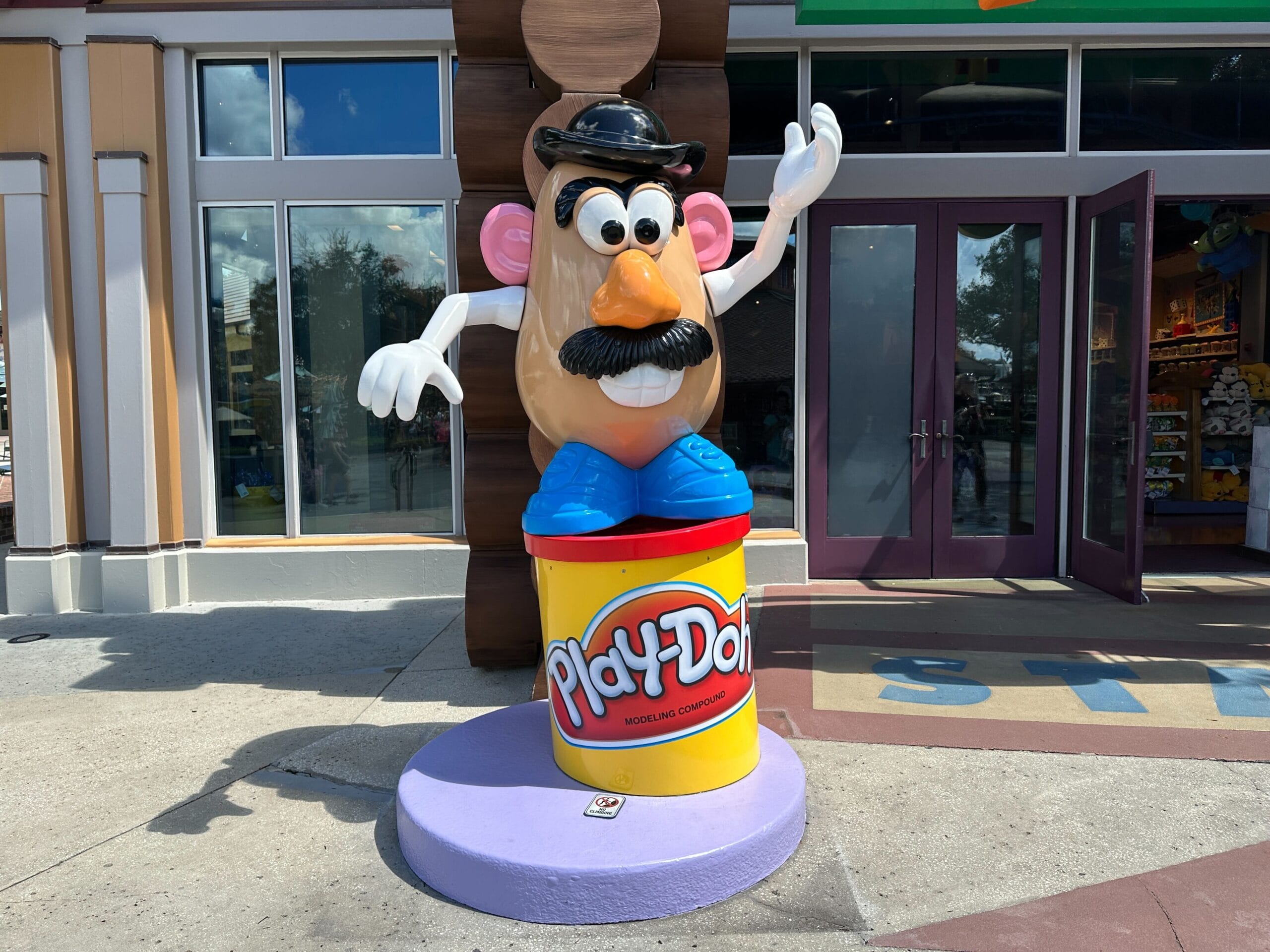Mr. Potato Head statue stands on a Play-Doh can outside a store, waving with one hand raised.