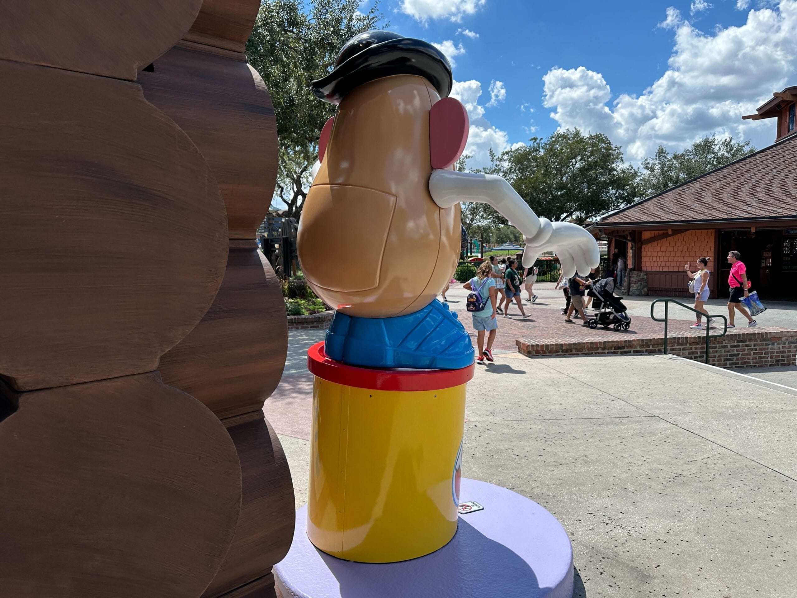 A statue of Mr. Potato Head faces away from the camera near a log cabin and people walking outside.