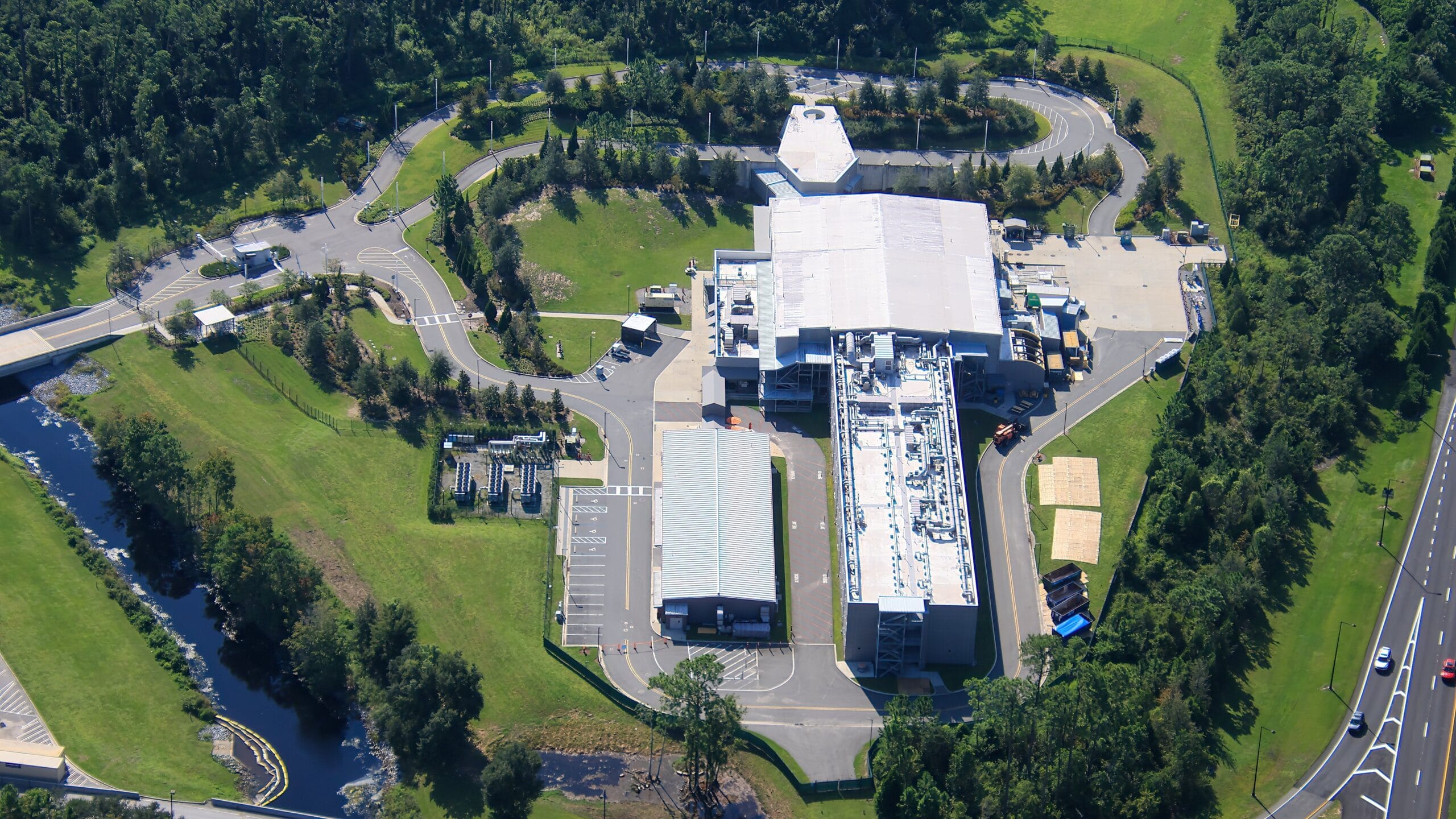 Aerial view of a large industrial complex surrounded by trees and roads on a sunny day.