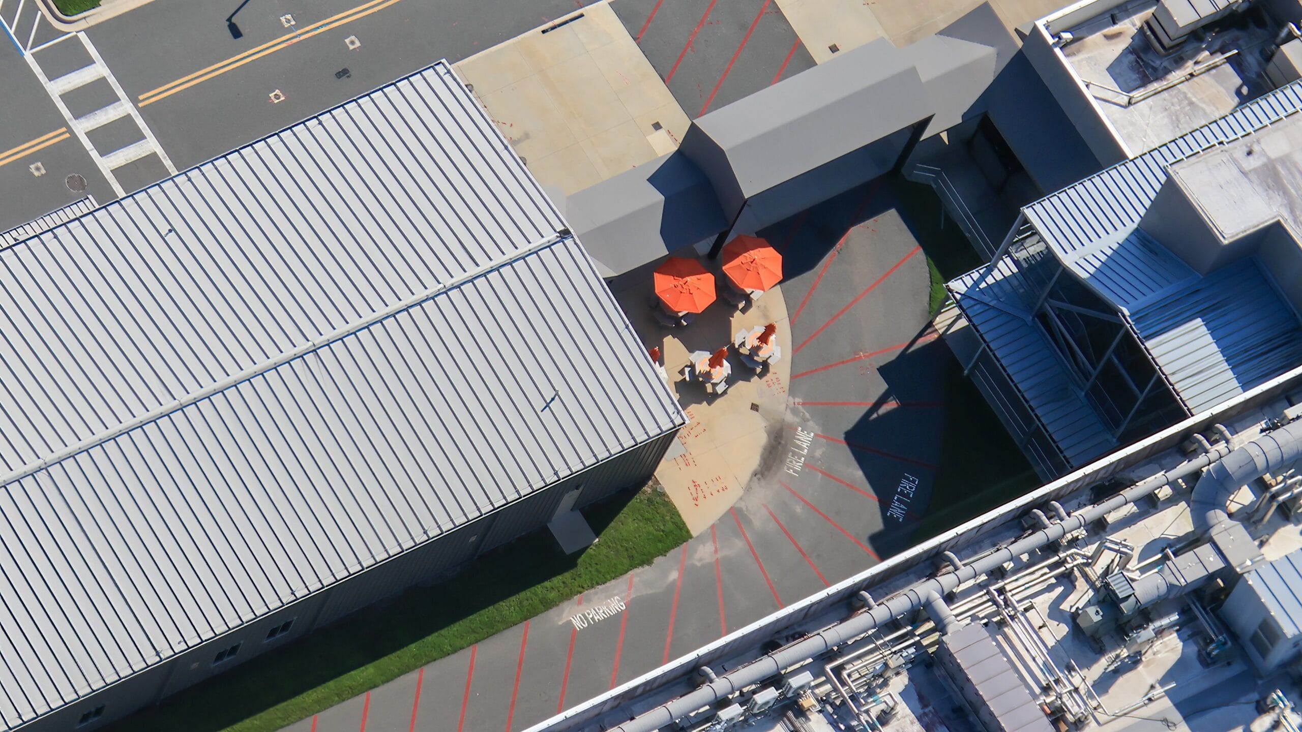 Aerial view of people sitting at tables with orange umbrellas between industrial buildings with gray roofs.