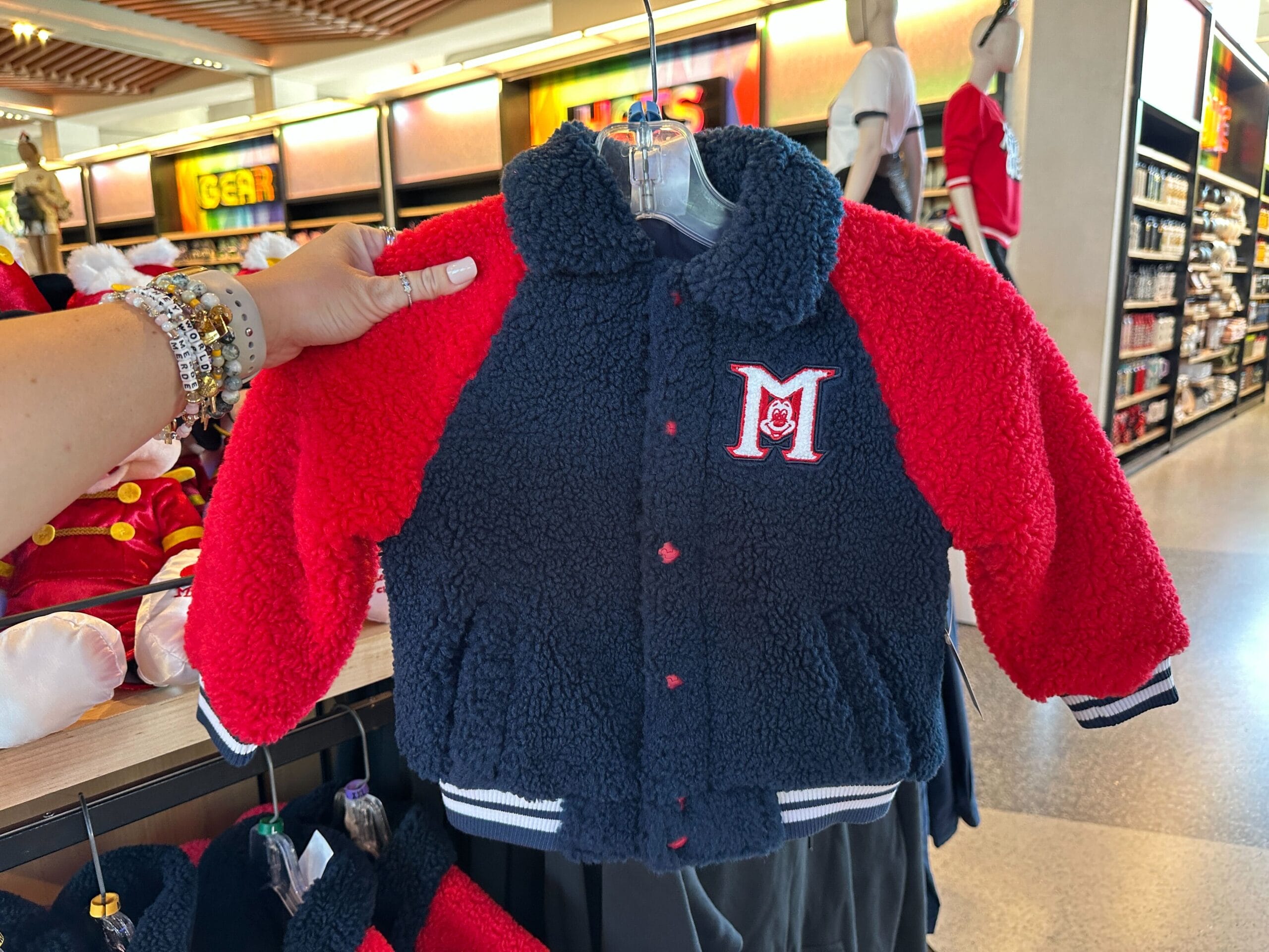 A hand displays a fuzzy red and navy jacket with an "M" patch from the Mickey Mouse Club 70th Collection in a Disney park shop.