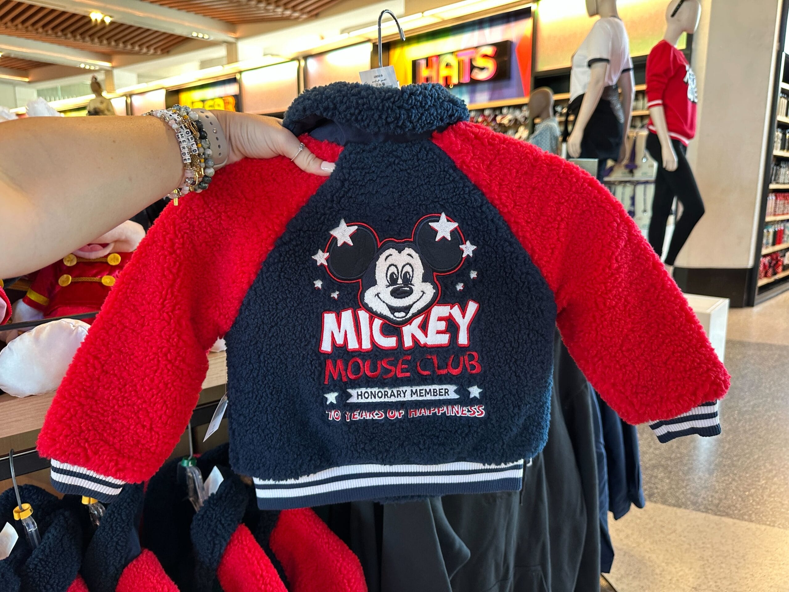 In a Disney Park store, a hand displays a red and navy 70th Mickey Mouse Club kids jacket with “Honorary Member” on it.