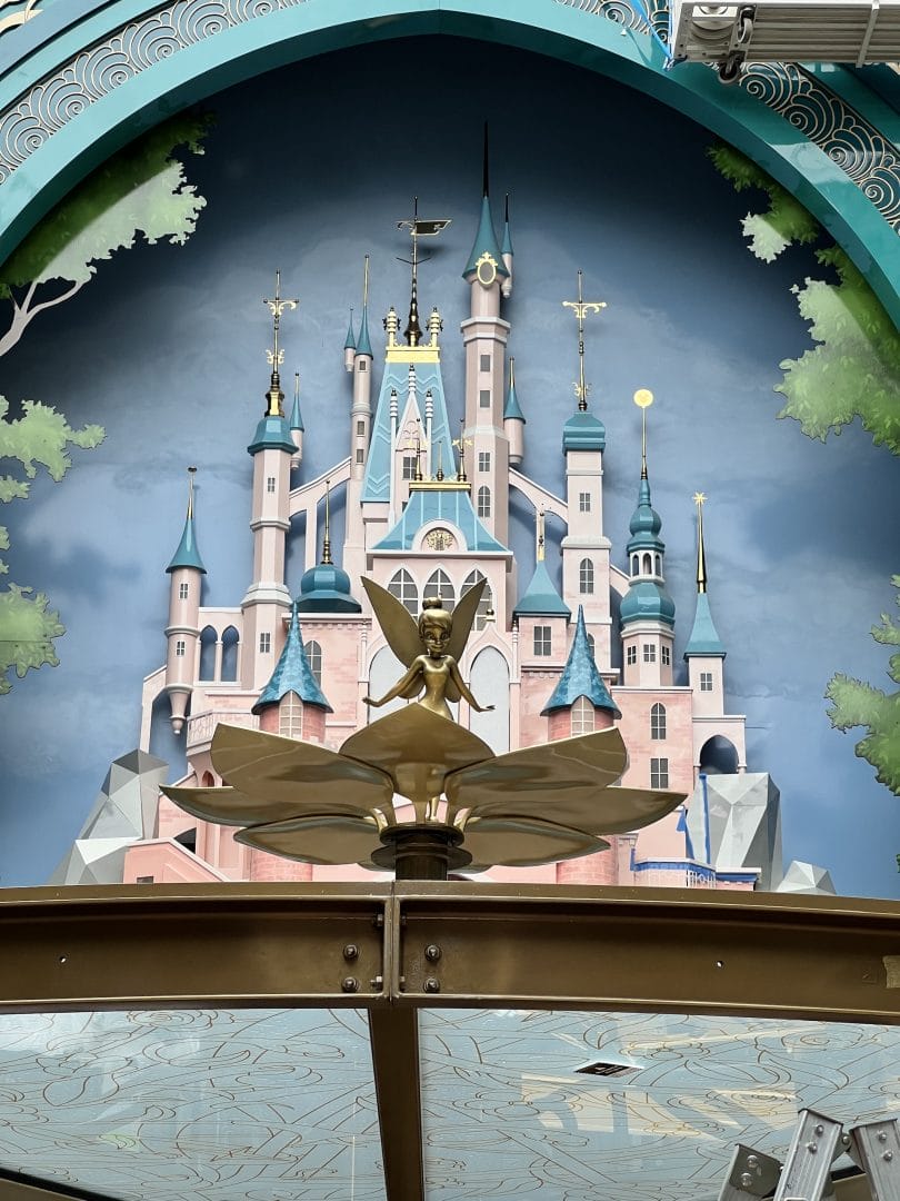 A gold Tinker Bell statue stands before a painted fairytale castle with blue spires, resembling Disneyland’s Sleeping Beauty Castle.