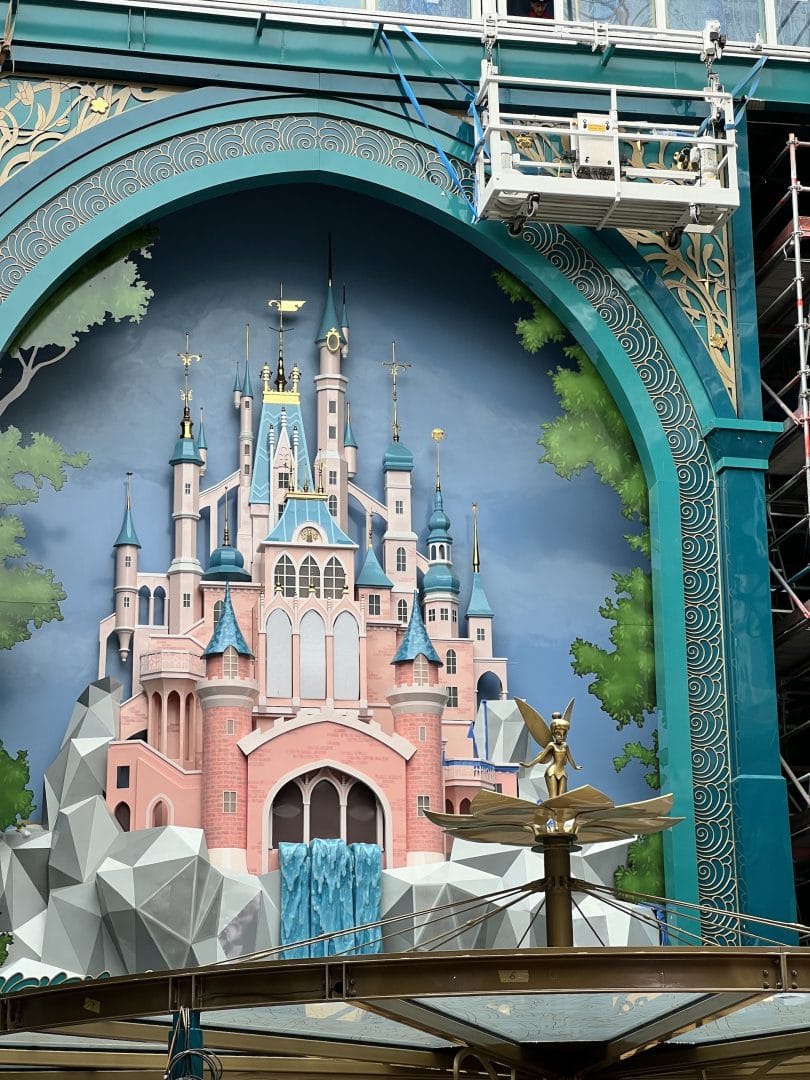 A mural of a pink and blue castle, reminiscent of Sleeping Beauty Castle, framed by arches with a golden Tinker Bell statue in front.