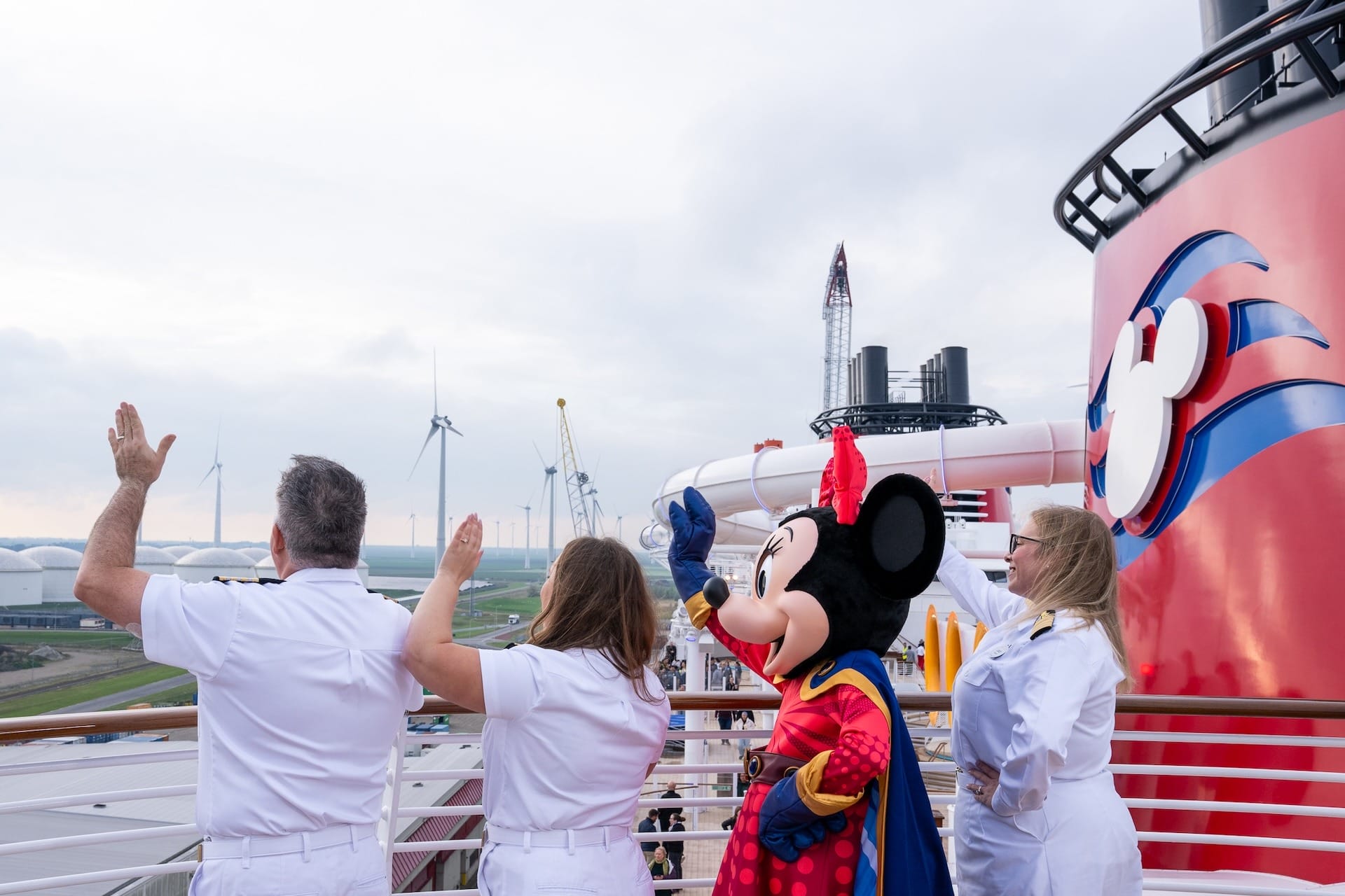 Three Disney cruise crew and Minnie Mouse wave on deck; wind turbines and cranes behind, not a Disney castle like in the parks.