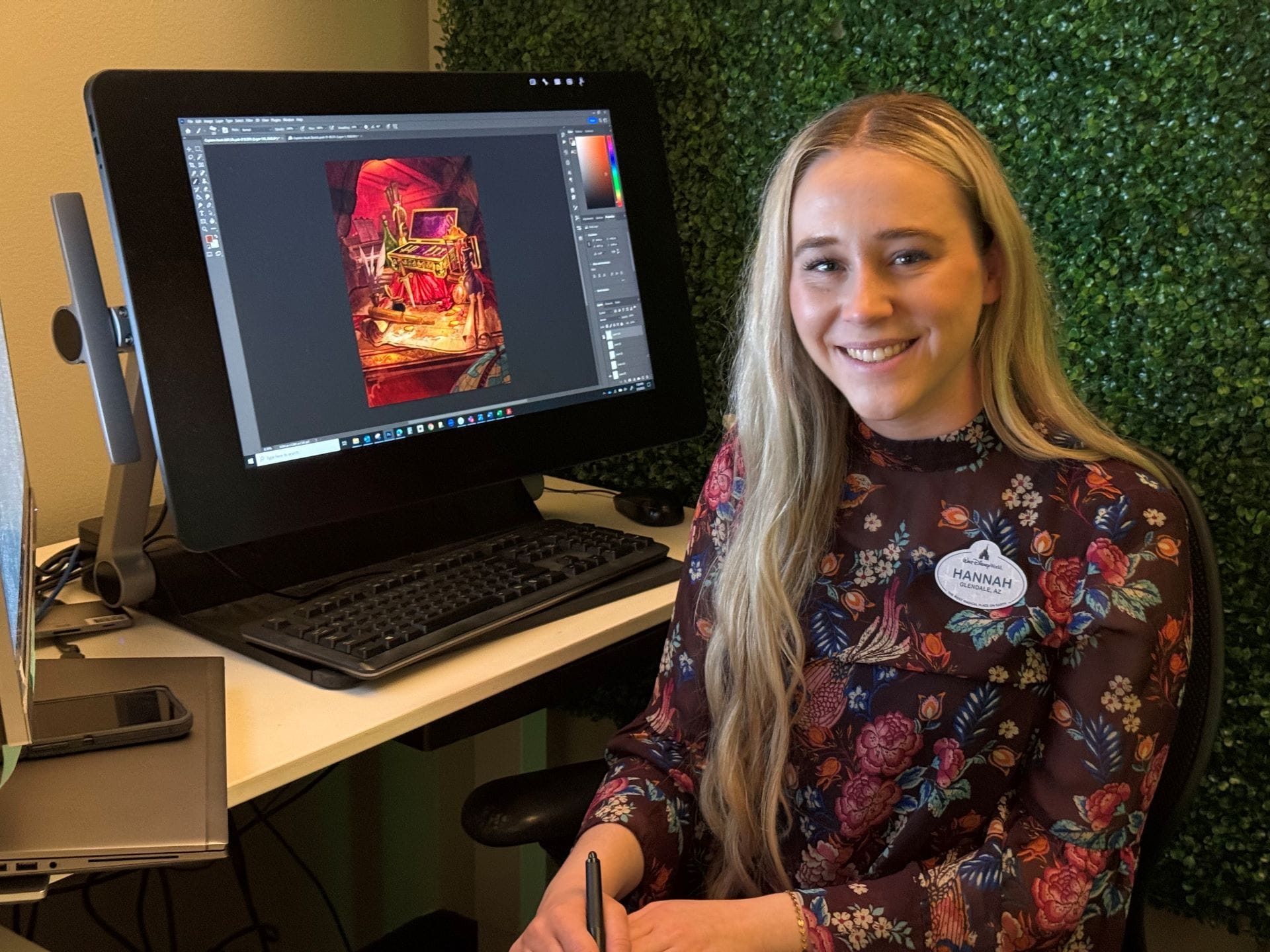 Woman with long blonde hair smiles at desk, Disney Destiny art on monitor, holding stylus pen; no castle visible for park comparison.
