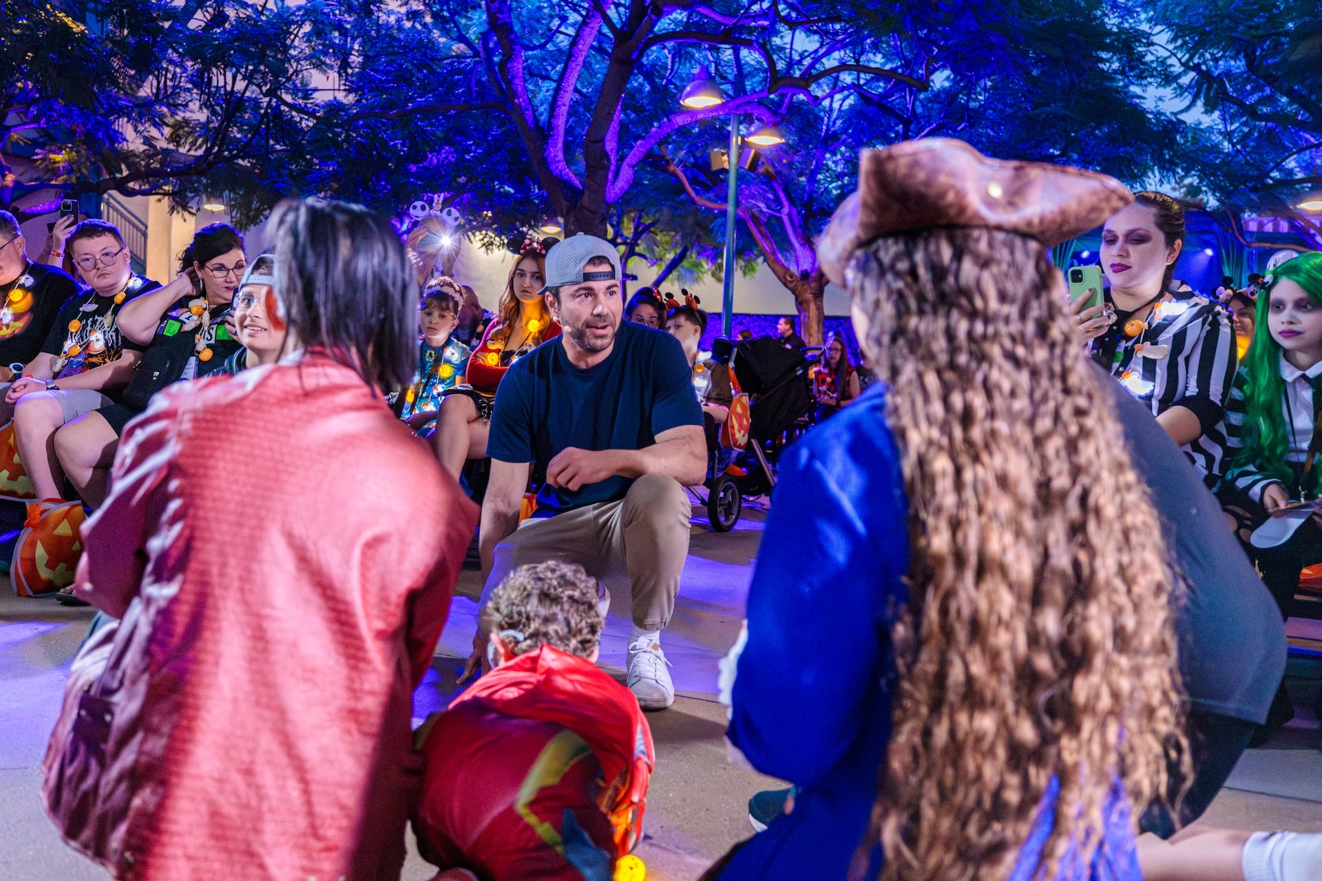 A man talks with costumed guests at a Disney park event under blue-lit trees at night, no castle seen to compare.