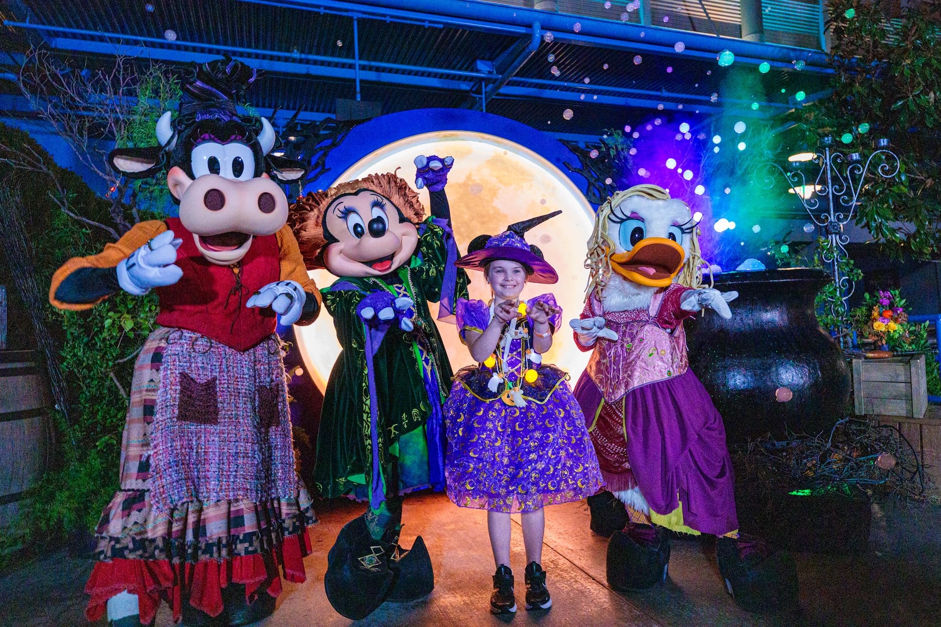 A child in a witch costume poses with Disney characters in Halloween outfits at Disneyland Resort, with Sleeping Beauty Castle nearby.