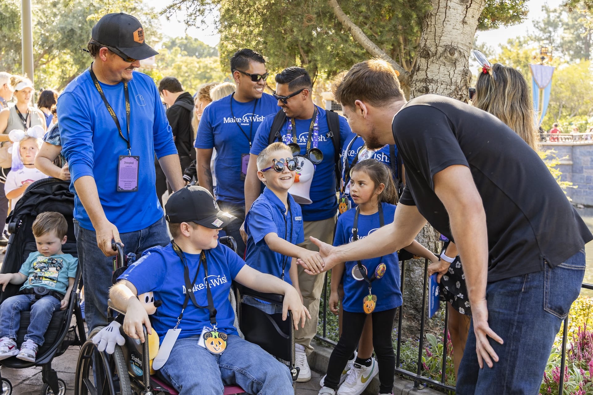 A man shakes hands with a smiling child in a wheelchair among a group in blue Make-A-Wish shirts at Disneyland Resort Anaheim.