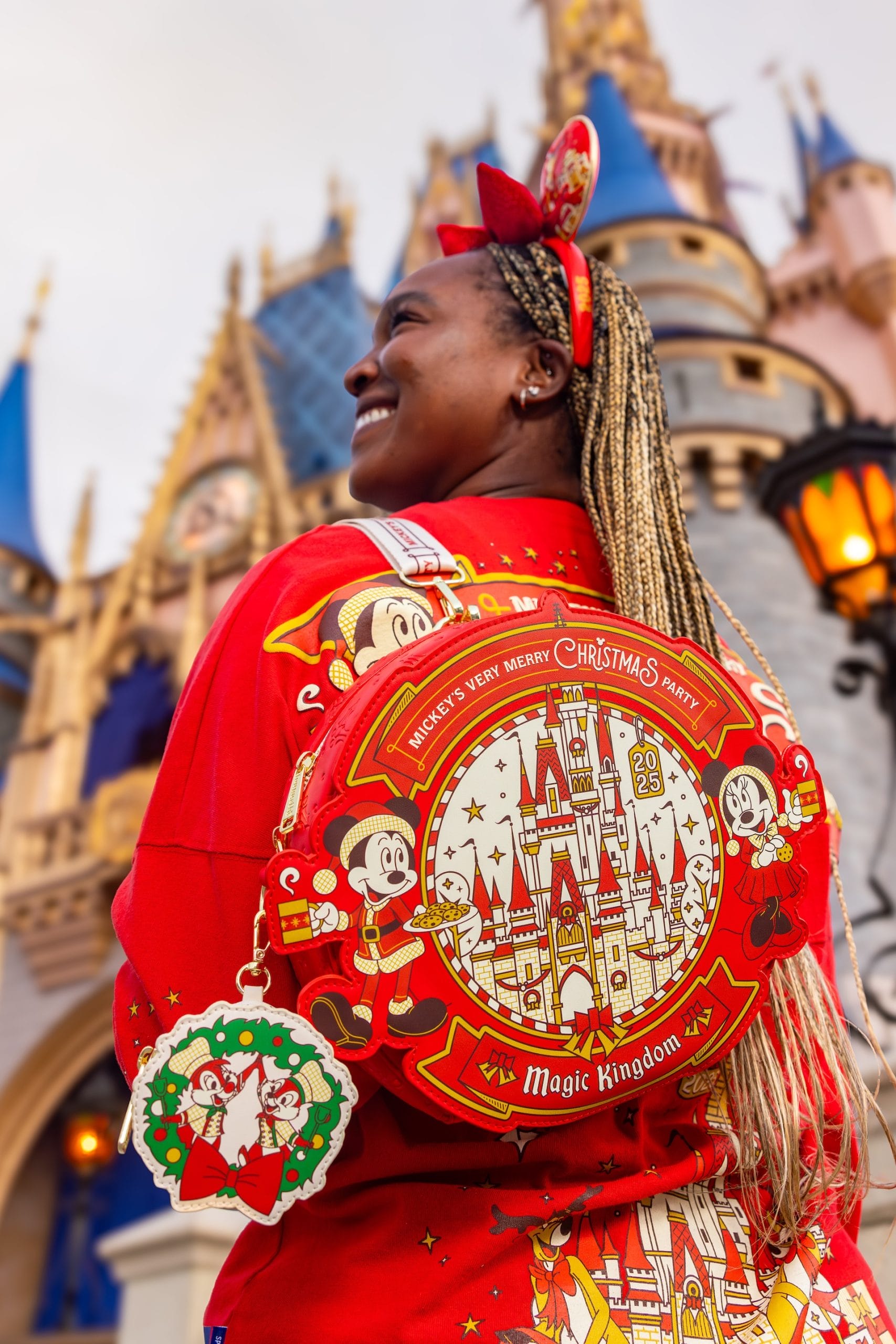 A woman in red festive attire poses at Magic Kingdom, with Cinderella Castle behind her, during a 2025 merchandise preview event.