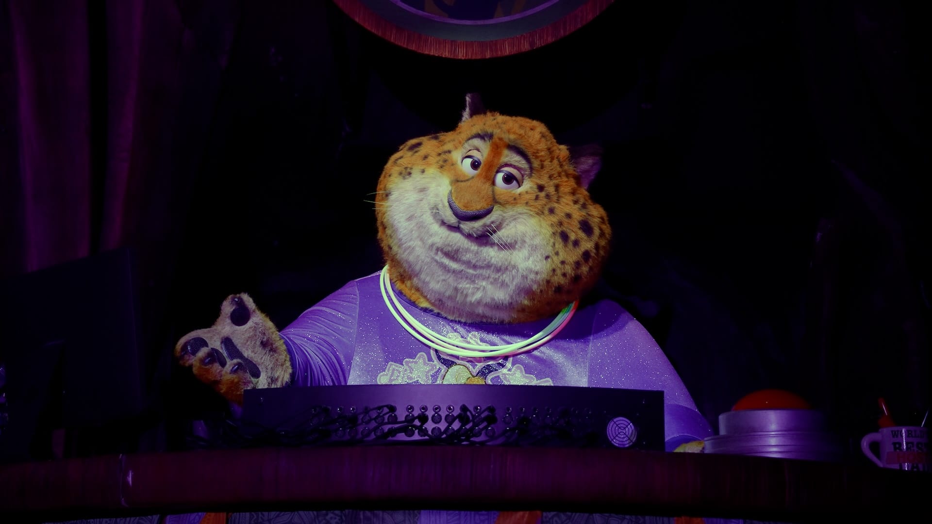 A costumed Zootopia cheetah in a purple outfit and glow necklaces entertains on a dark Disney Parks stage, no castle in view.