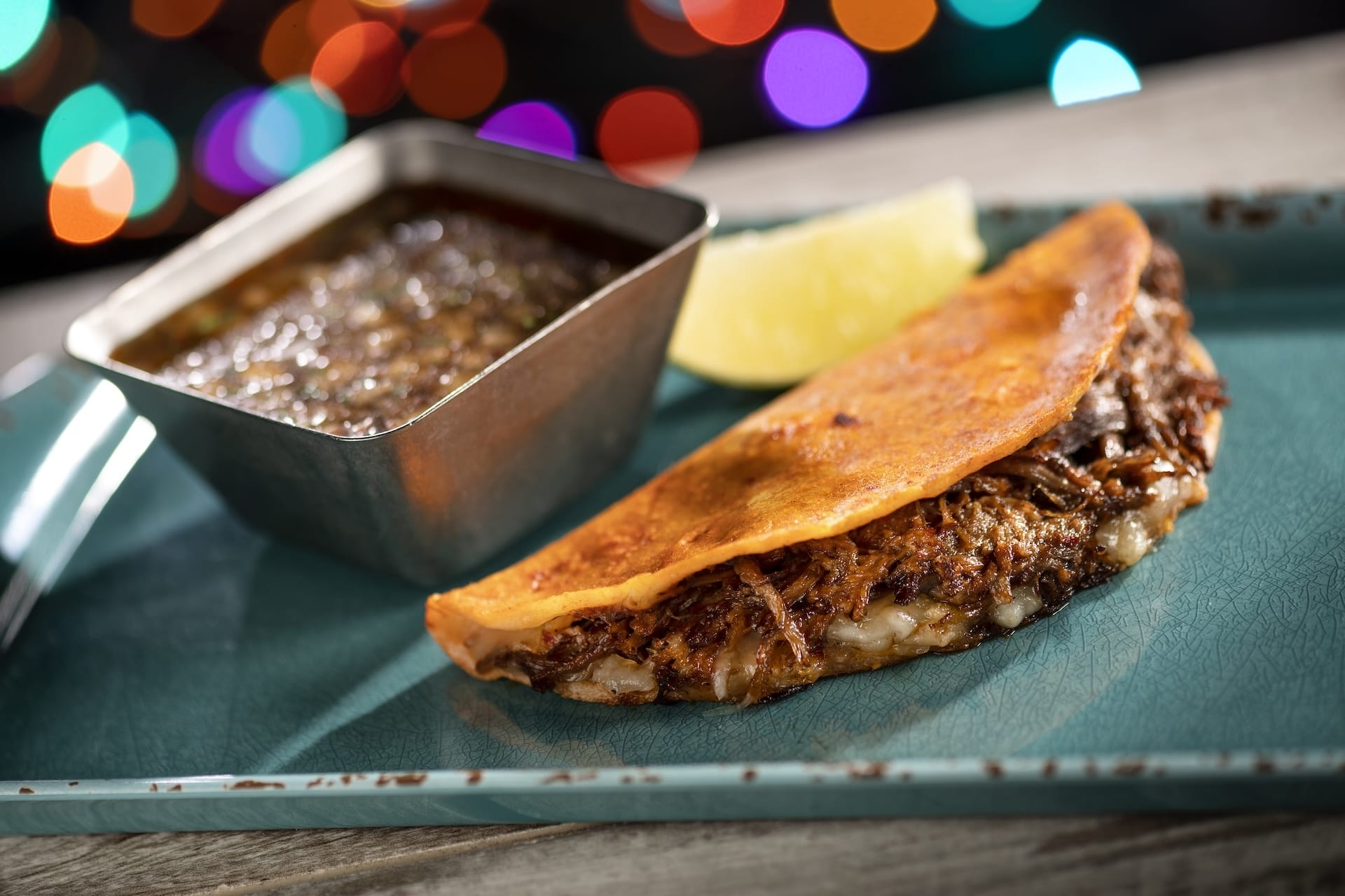 A Disney Jollywood Nights birria taco with gooey cheese, sauce, and lime on a teal plate, a festive new treat at Walt Disney World.