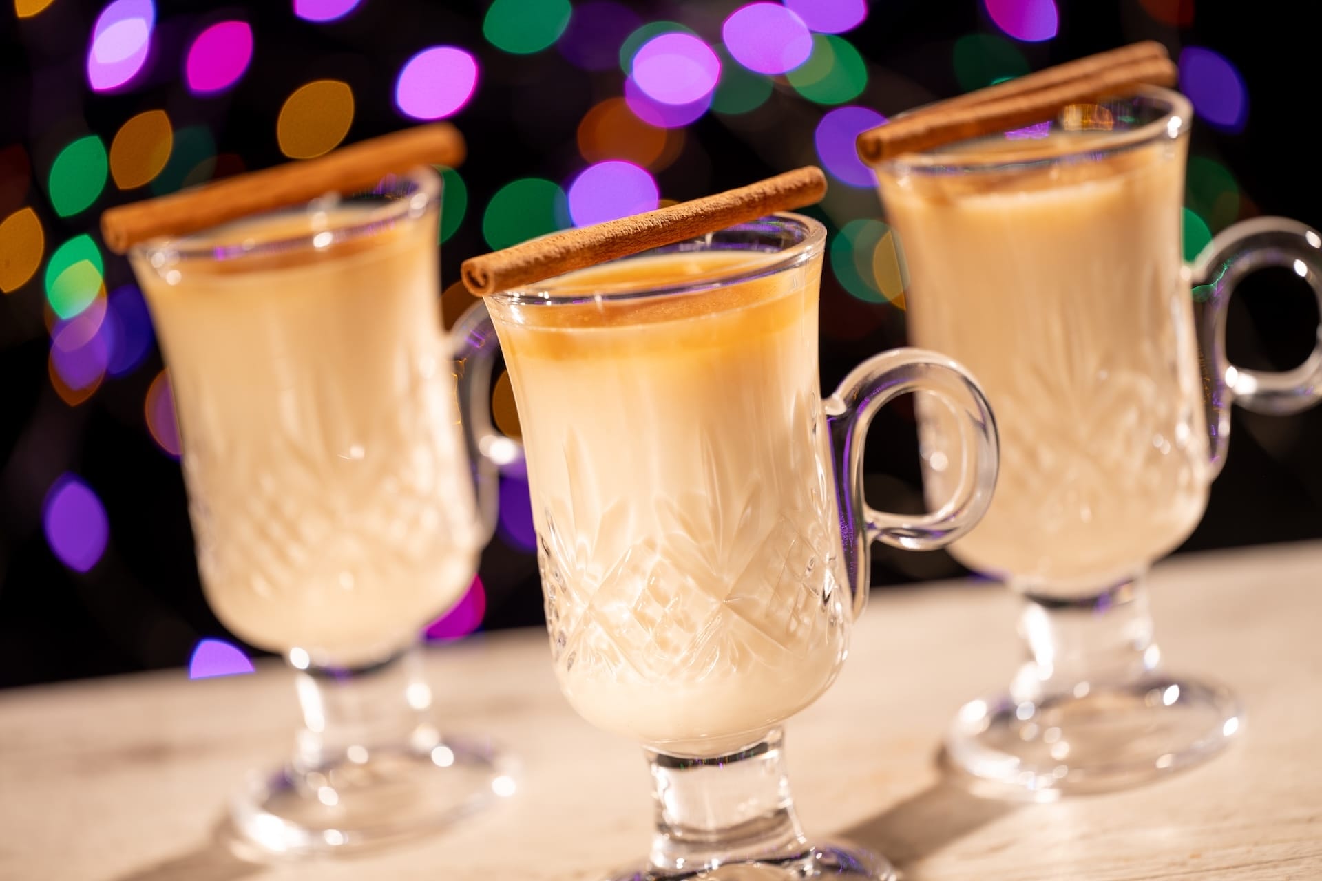 Three glass mugs of creamy eggnog, a festive new treat, with cinnamon sticks and twinkling holiday lights evoke Main Street U.S.A.