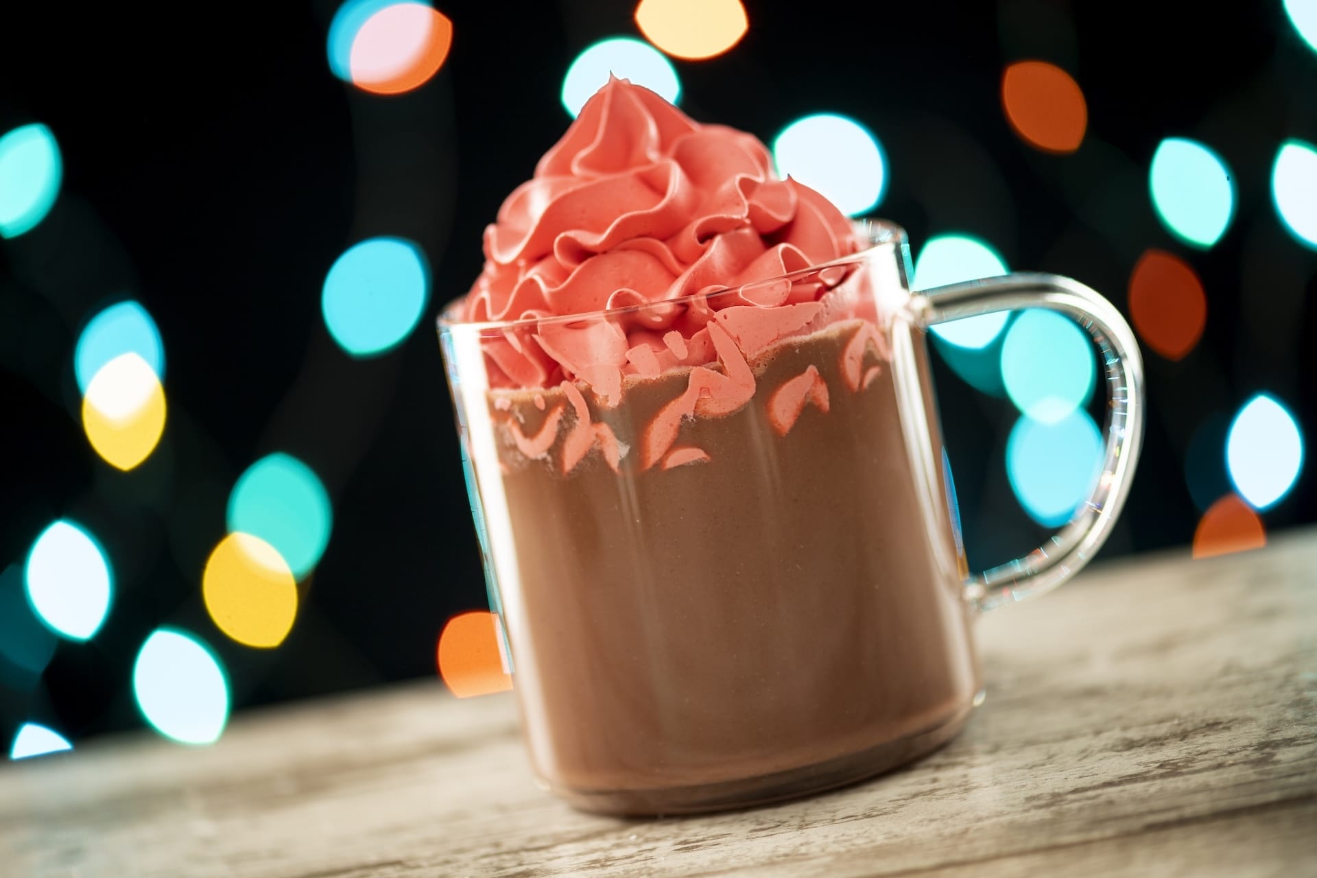 A glass mug of hot chocolate with pink whipped cream glows amid vibrant lights at Disney Jollywood Nights 2025 in the parks.