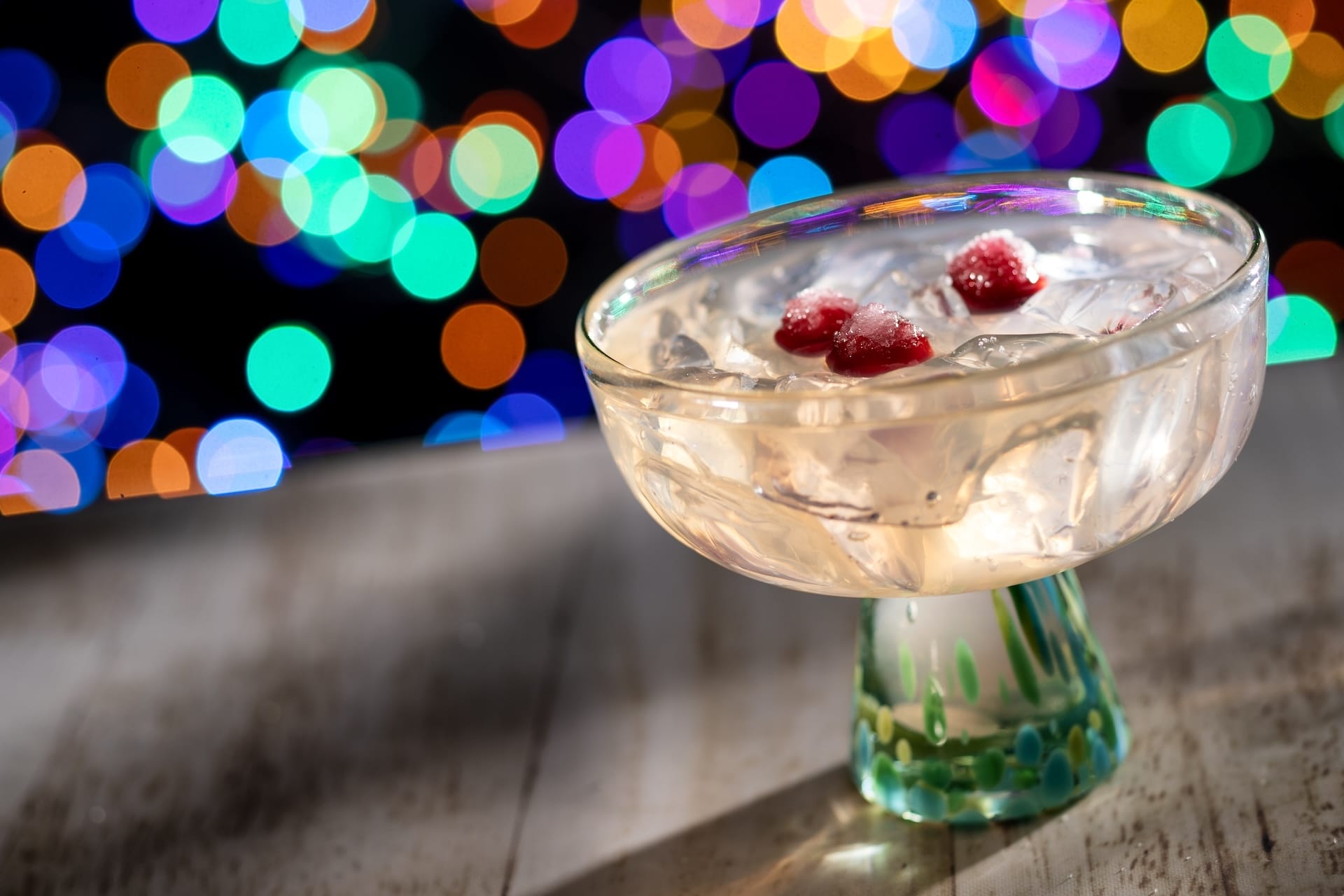 A festive cocktail glass with ice and red berries, ideal for Disney Jollywood Nights, shines before vibrant, dreamy park lights.