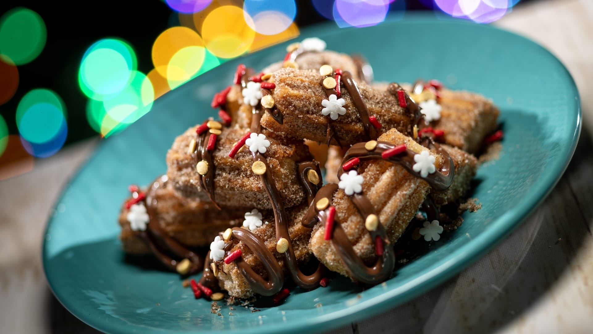 Churros with chocolate drizzle and sugar snowflakes on a teal plate, festive lights in the background at Disney Jollywood Nights.