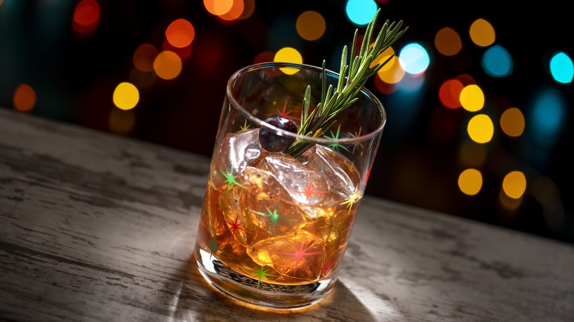 A festive cocktail with ice, cherry, and rosemary on a table at Disney Jollywood Nights, with vibrant park lights in the background.