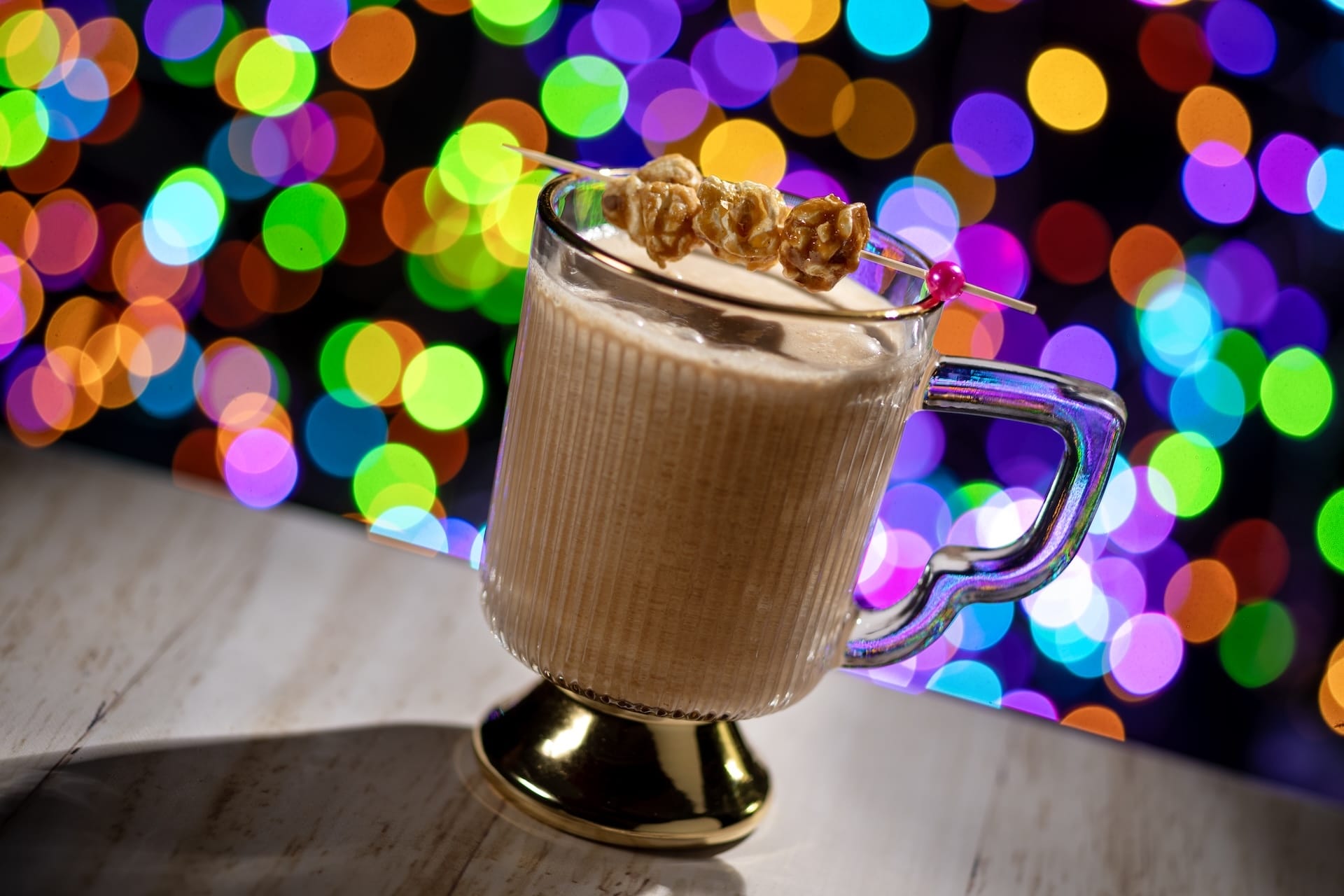 A creamy drink garnished with popcorn sits before vibrant Jollywood Nights lights at Disney, no castle but pure park magic.