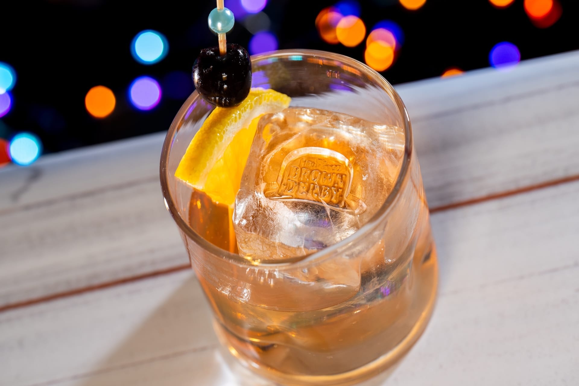 A cocktail with a large ice cube, orange slice, and cherry gleams under Disney Jollywood Nights' festive park lights. No castle in view.