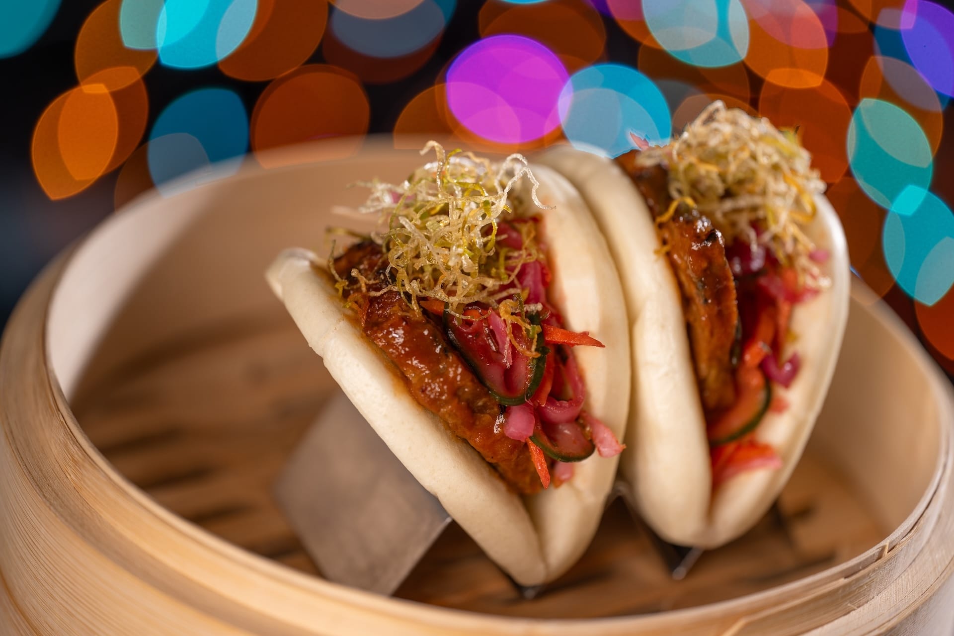 Bao buns stuffed with veggies and meat in a bamboo steamer at Disney Jollywood Nights, vibrant bokeh lights glowing in the background.