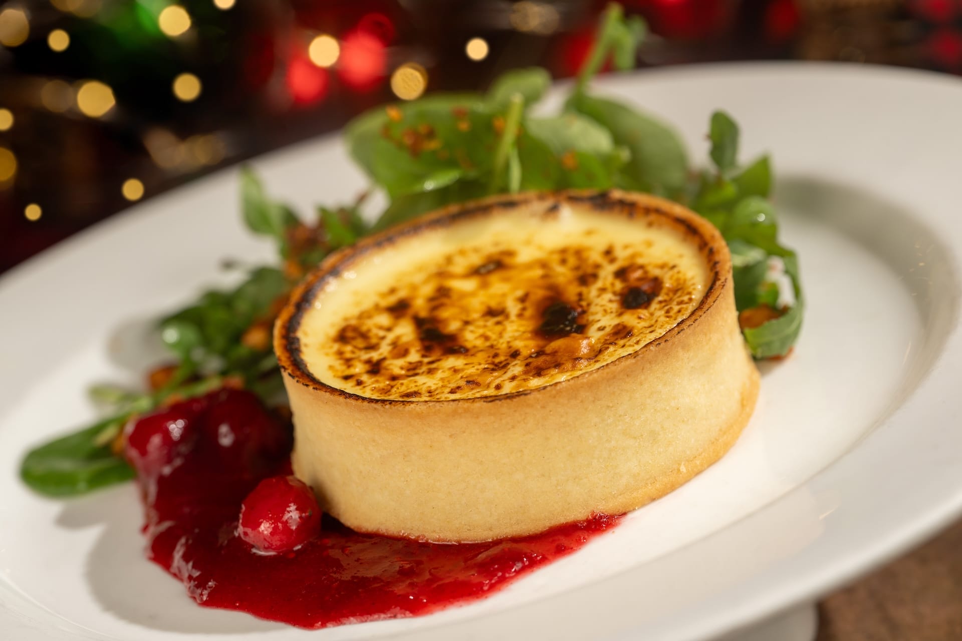 A golden-topped holiday tart with salad greens and berry sauce, plated elegantly like a Disney park dessert, on white china.