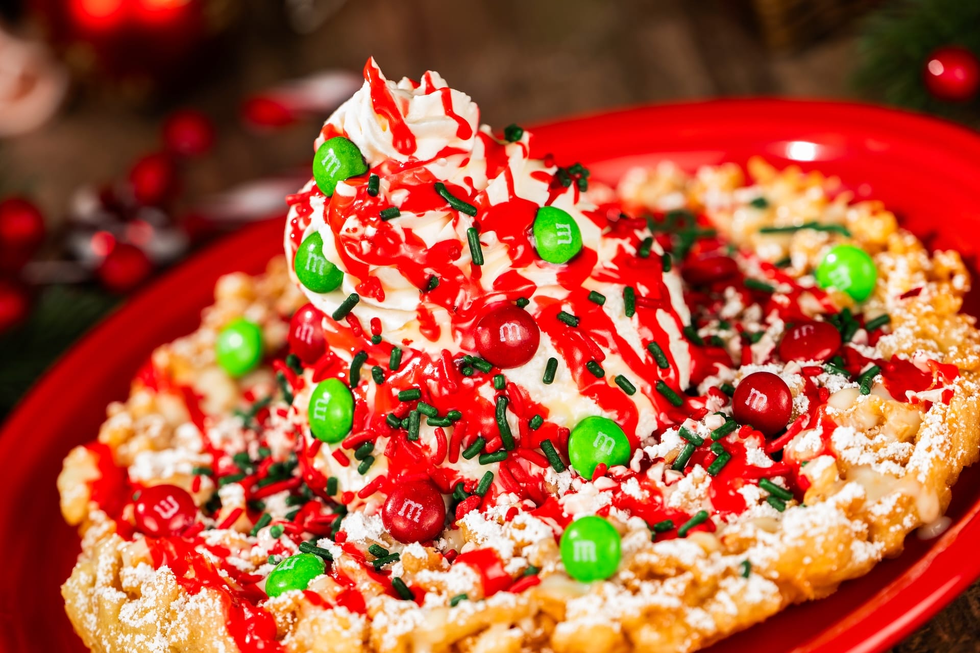 Holiday funnel cake at a Disney park, topped with whipped cream, red syrup, candies & sprinkles on a festive red plate.