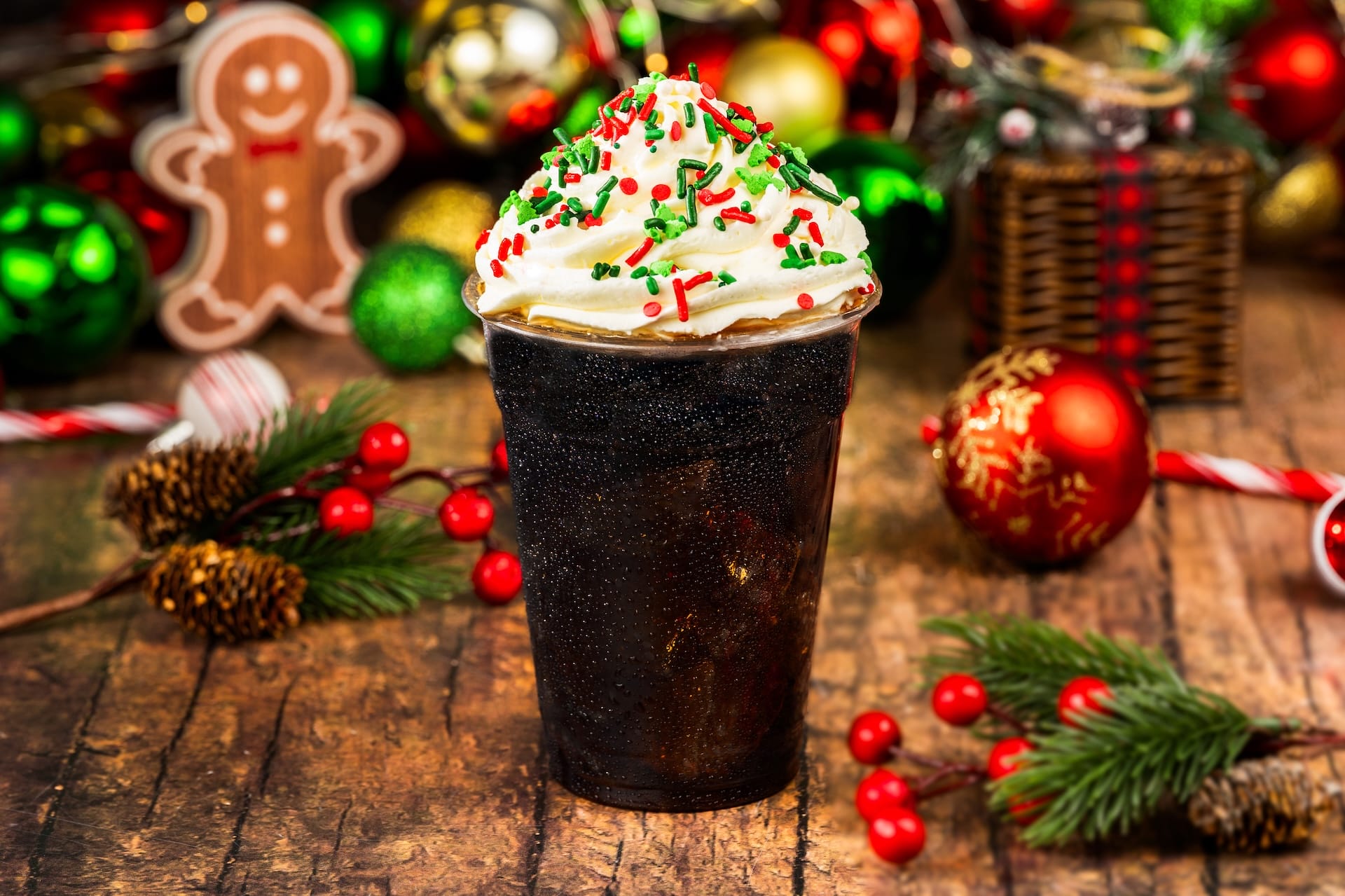 A festive holiday cola with whipped cream and sprinkles, surrounded by Disney-themed Christmas decor at a park during the holidays.