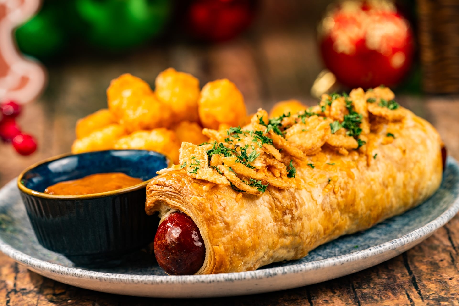 A festive sausage roll in flaky pastry, crispy onions on top, with tater tots and dipping sauce like a Disney Parks holiday treat.