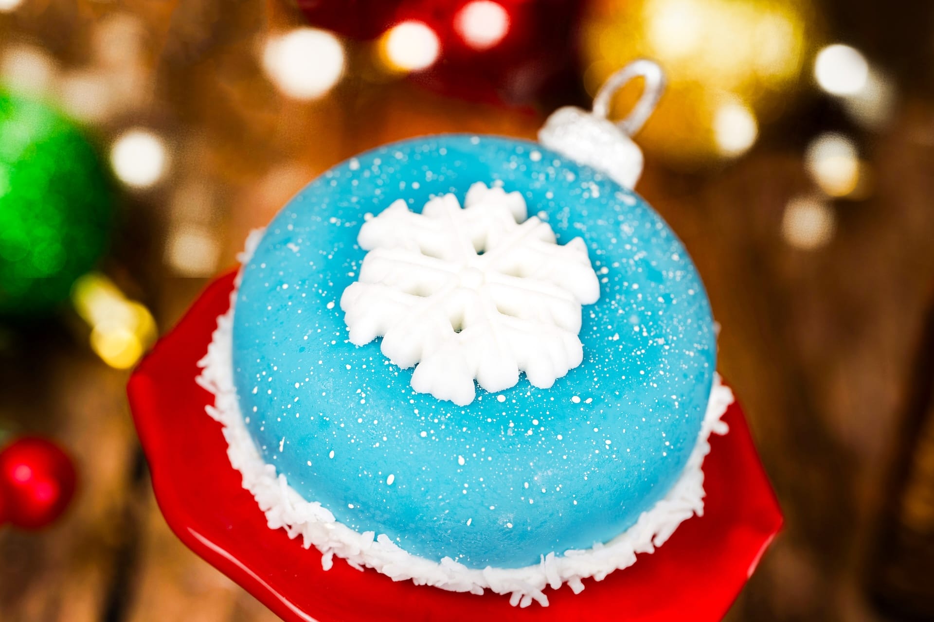A festive blue ornament-shaped cake with a white snowflake and coconut edges sits on a Holiday Menu red stand at a Disney park.