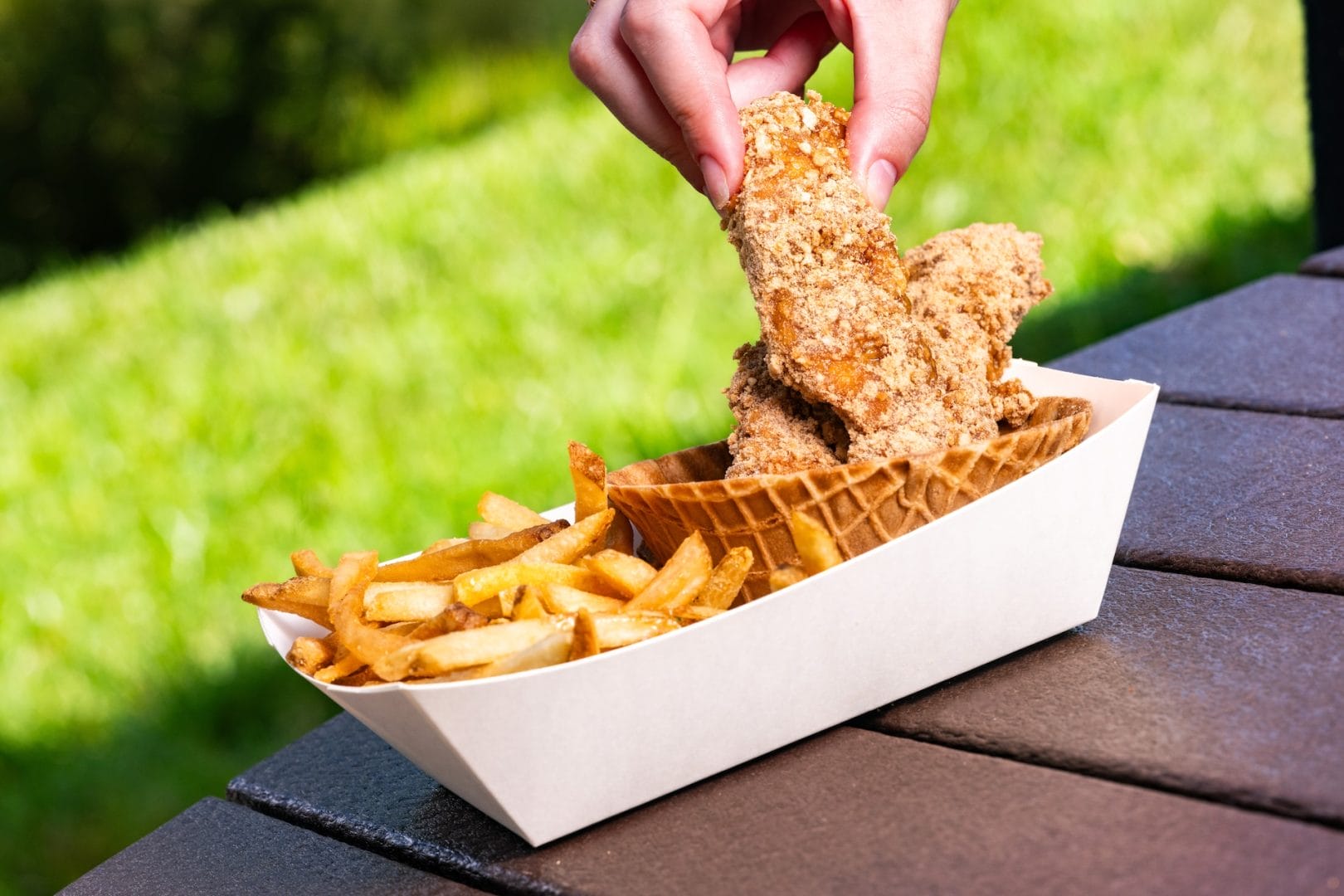 A hand picks up a Disney World chicken strip from a tray with fries and a waffle bowl on an outdoor table.