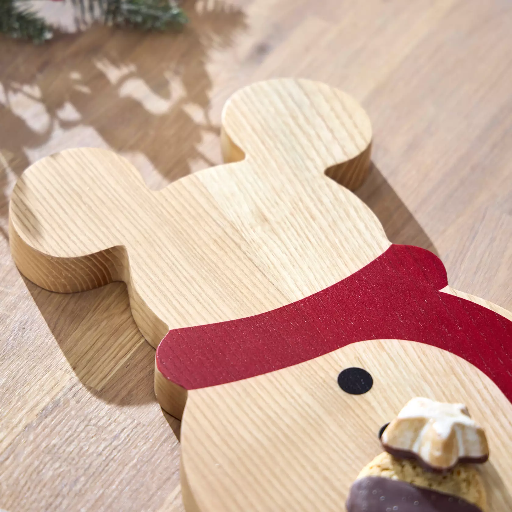 Wooden cutting board shaped like a Mickey snowman wearing a red scarf, displayed with holiday treats on a table.