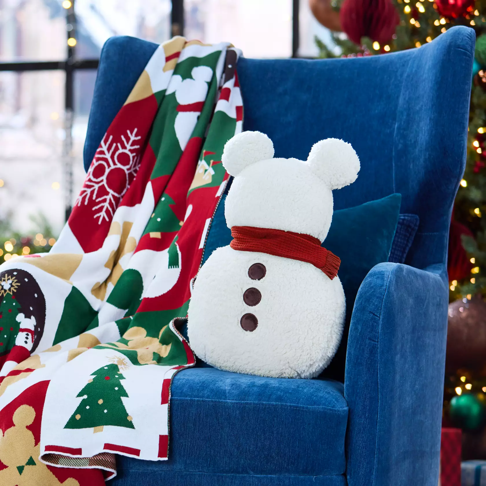 Mickey Snowman pillow on a blue chair next to a Disney Christmas throw blanket in front of a Christmas tree.