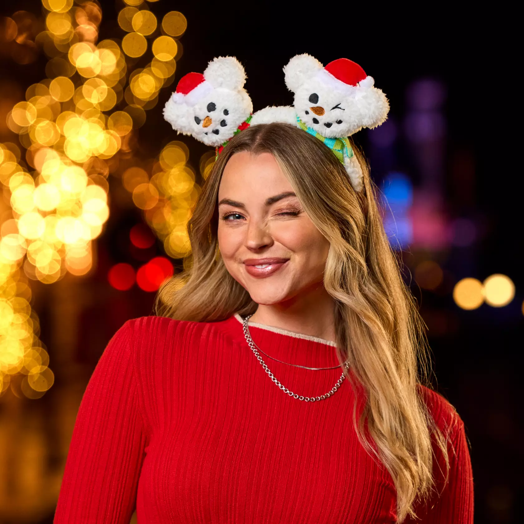 Woman in red sweater winking, wearing a plush Mickey Snowman headband. Blurred festive park lights sparkle behind her.