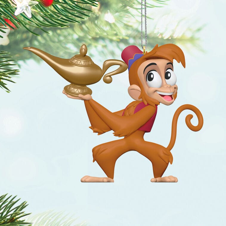 Cartoon monkey Disney ornament with magic lamp, like Hallmark at Disney Parks, hangs on pine tree amid festive park decor.