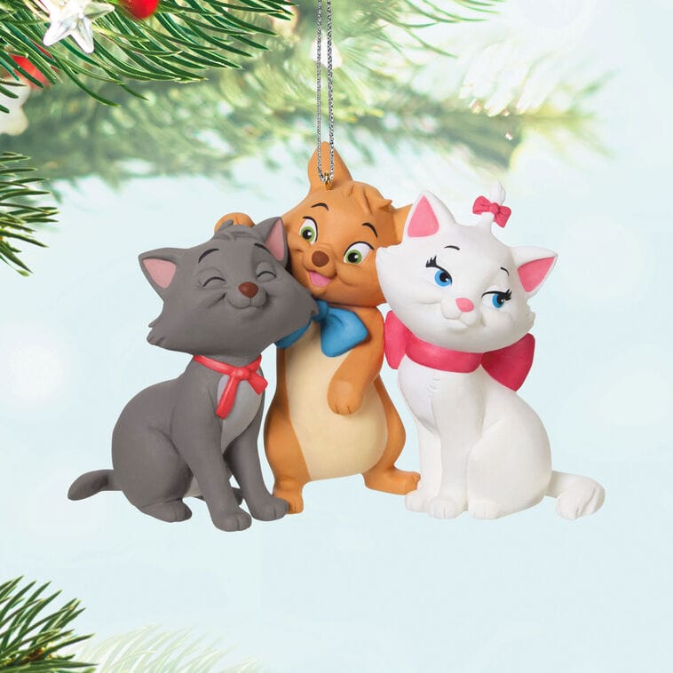 Three cartoon kittens with bows sit together as a Hallmark Christmas tree ornament, framed by branches—evoking Disney park charm.
