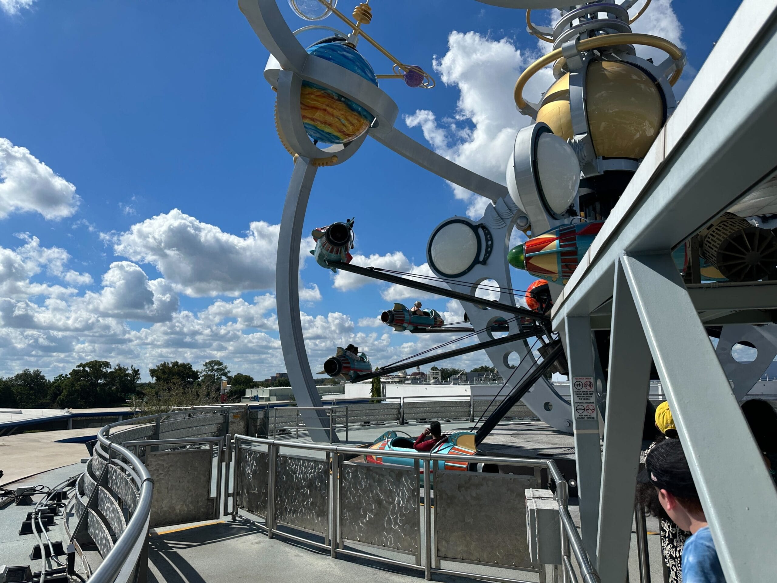 The Astro Orbiter rockets spin against a bright blue sky with fluffy clouds, just like at several Disney parks near their iconic castles.