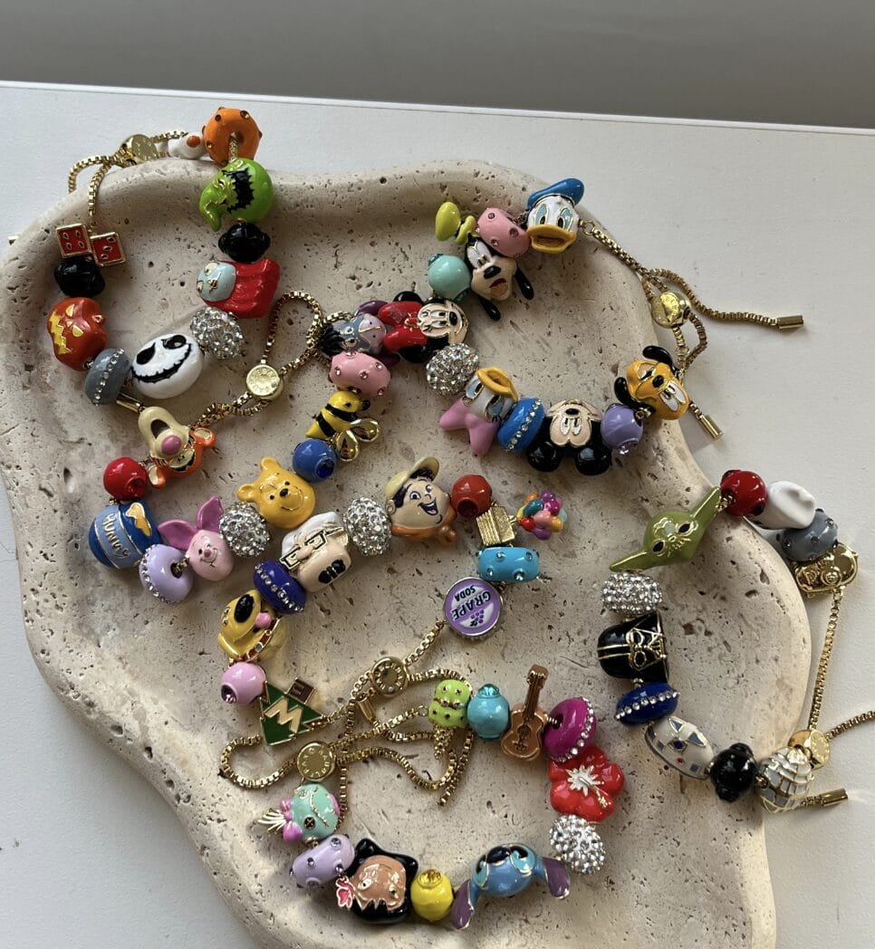 Three gold charm bracelets with vibrant Disney character beads and charms, reminiscent of Disneyland souvenir jewelry, on a speckled tray.