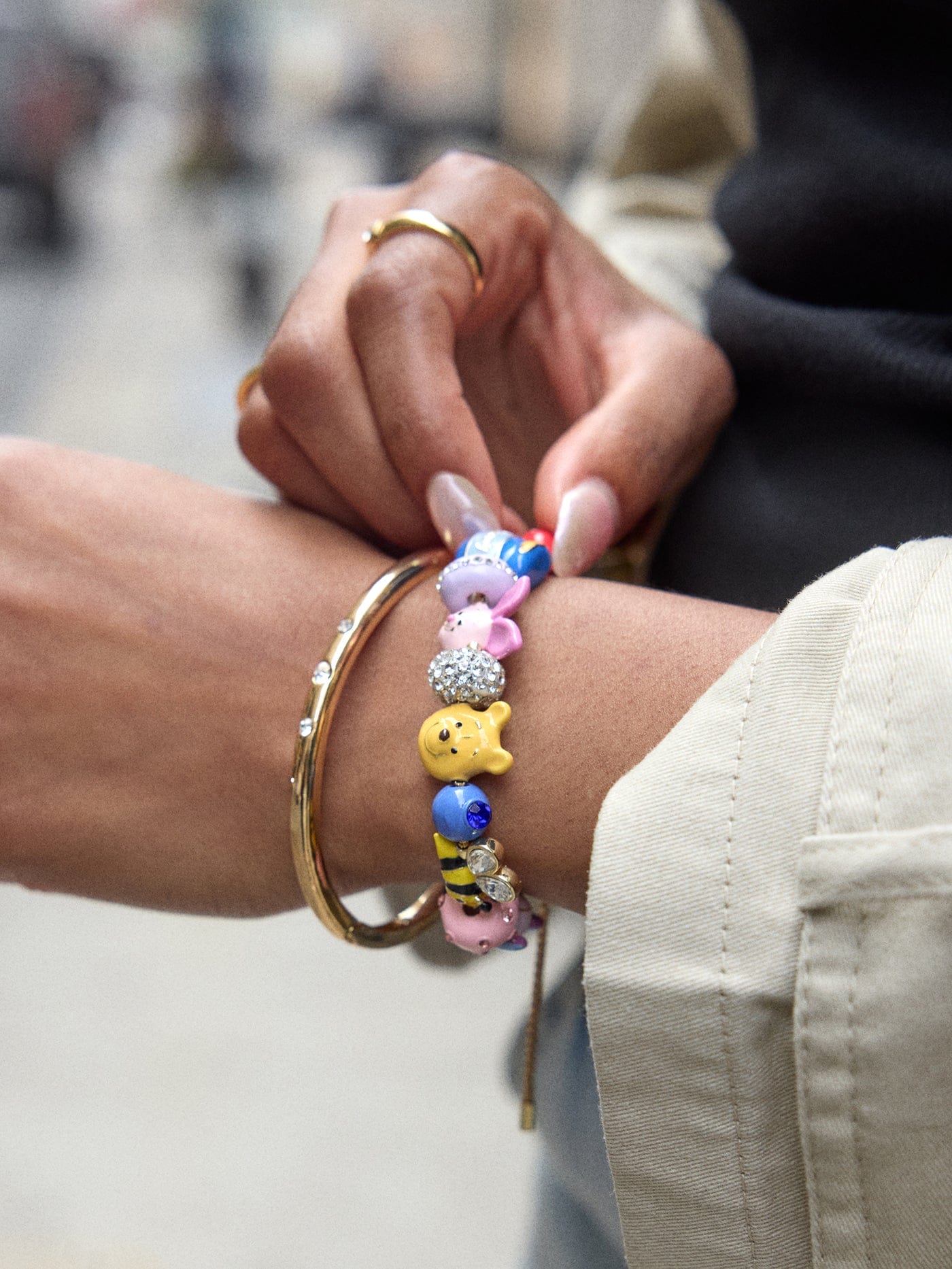A person at a Disney park wears gold bangles and a colorful animal-charm bracelet, holding their wrist in a magical setting.