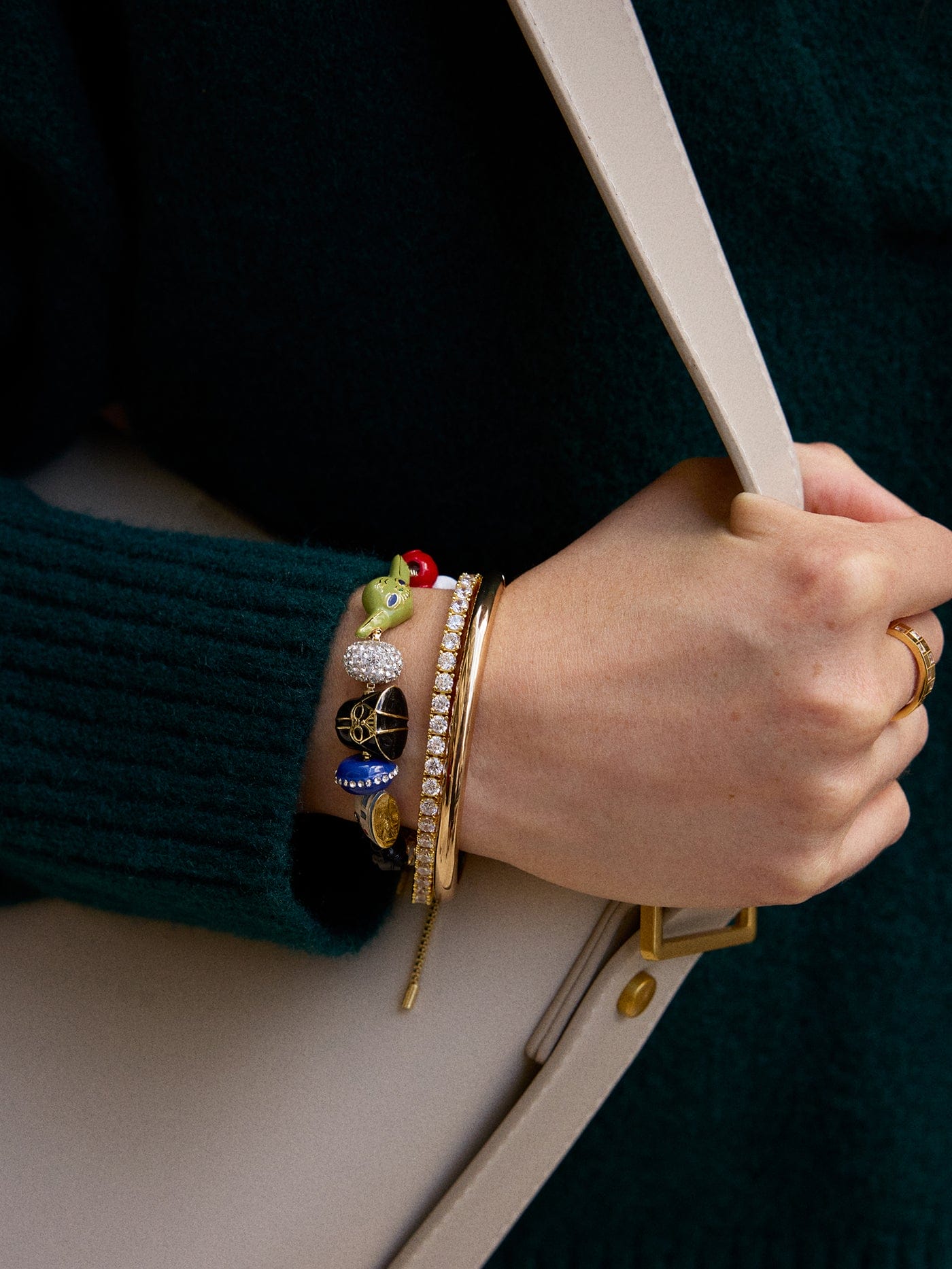 A hand with colorful Disney-themed charm bracelets and gold bangles holds a beige bag, the arm in a dark green sweater at the park.