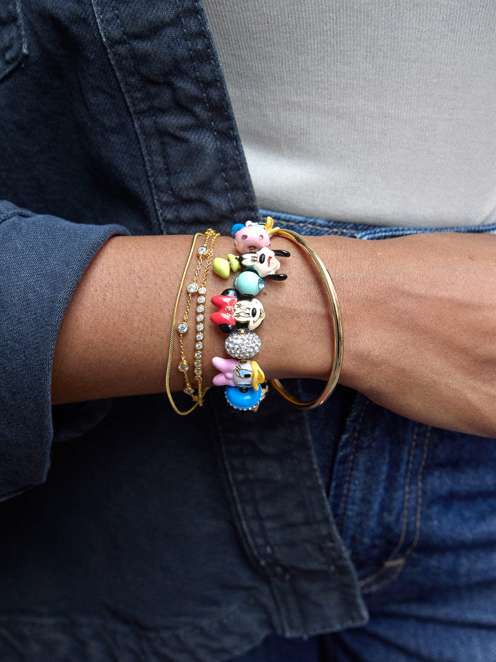 A person’s wrist with colorful charm bracelets, a gold bangle, and a chain bracelet, styled for a day at Disney in denim attire.