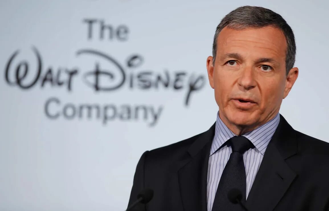 A man in a suit gives a daily recap at a podium with The Walt Disney Company logo in the background.